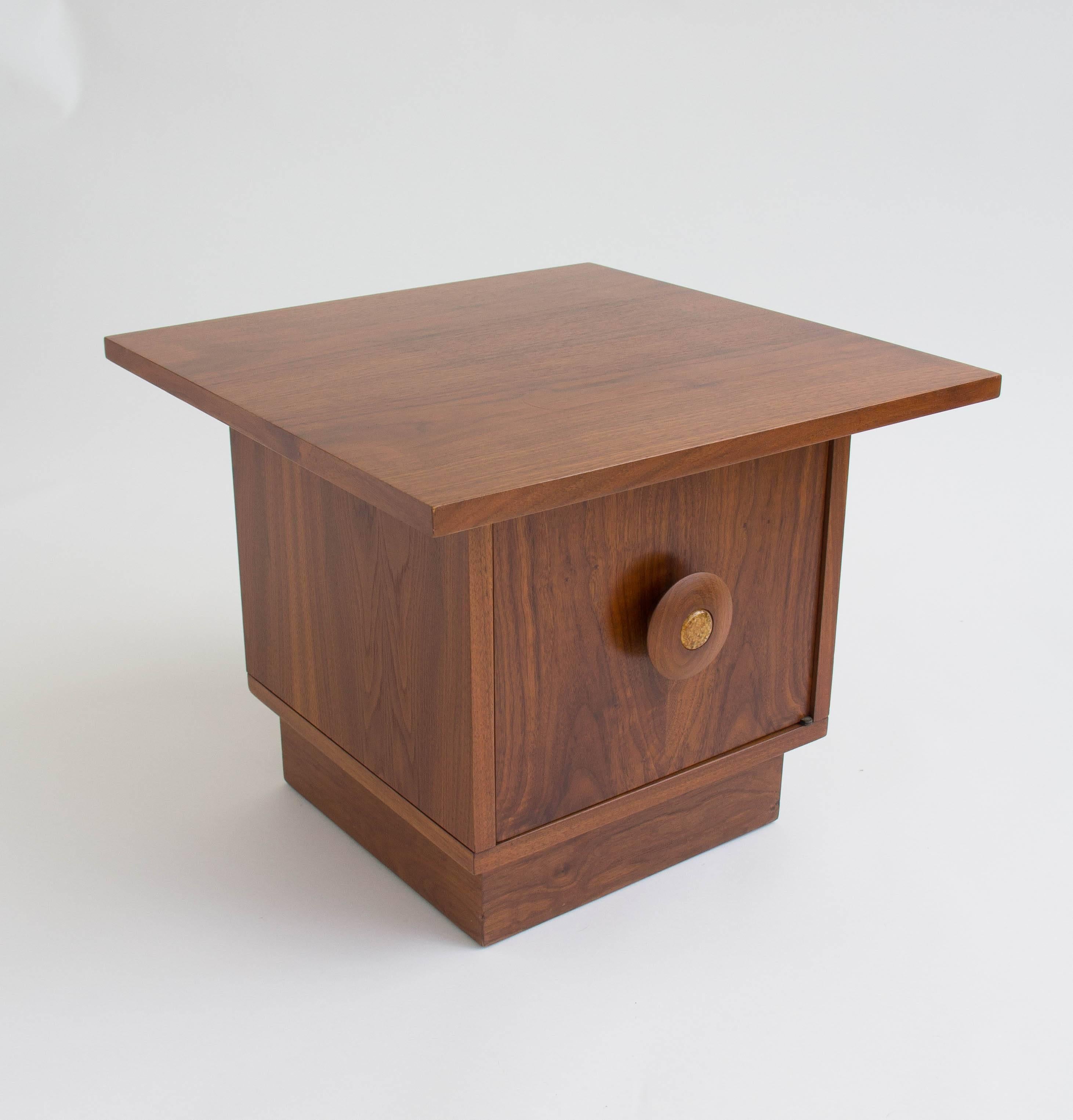 A pair of pedestal nightstands or low occasional tables in walnut by John Keal for Brown Saltman. Each piece has a square table top mounted on a pedestal with interior storage. The storage compartment opens with an oversized round knob, inlaid with