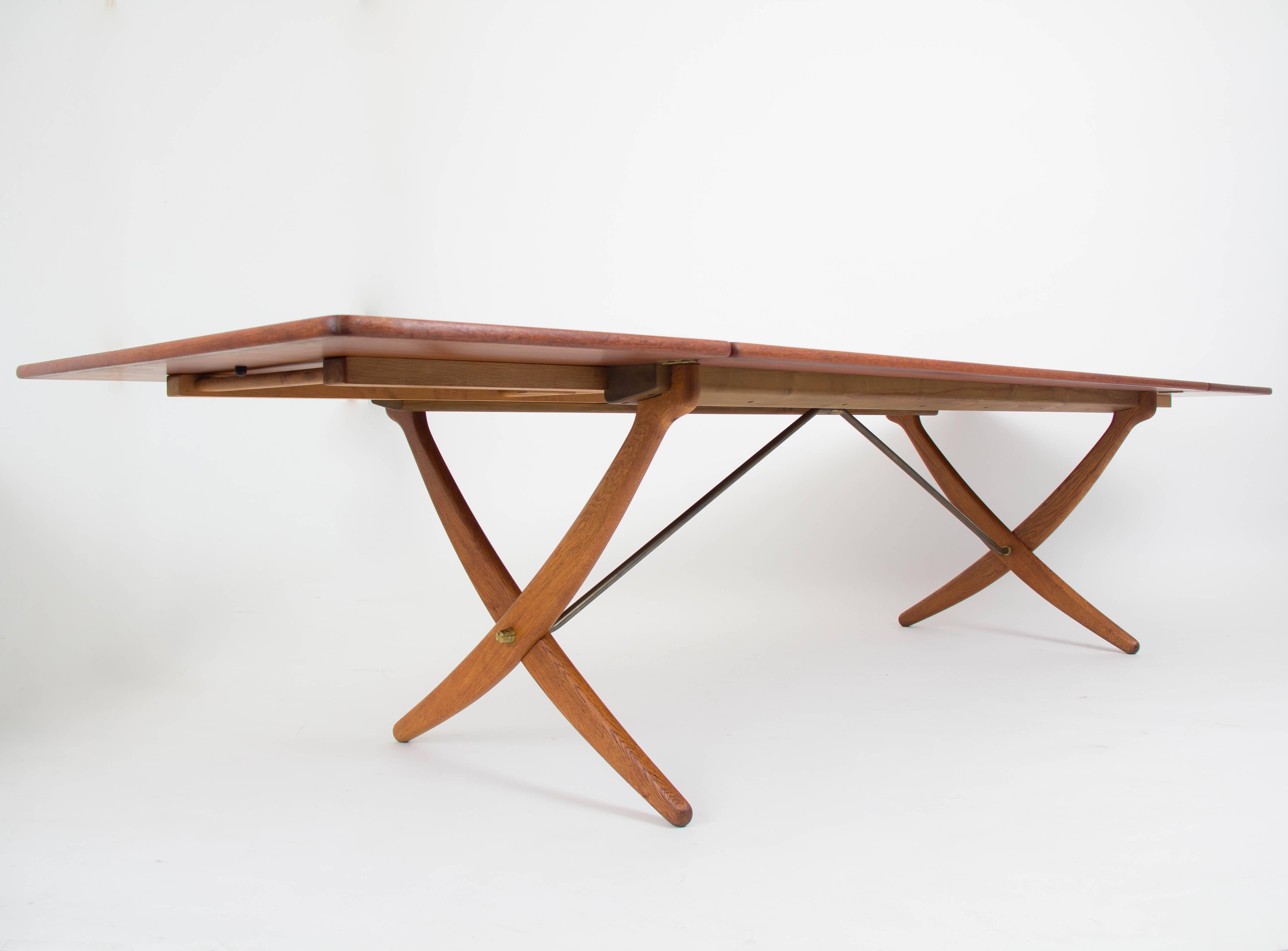 Hans Wegner’s AT-314 dining table by Andreas Tuck has two drop leaves and a distinctive crossed sabre legs.  The generous tabletop is teak, with edging in solid teak framing the top and leaves.  A wooden bar pulls out to support the leaves. The