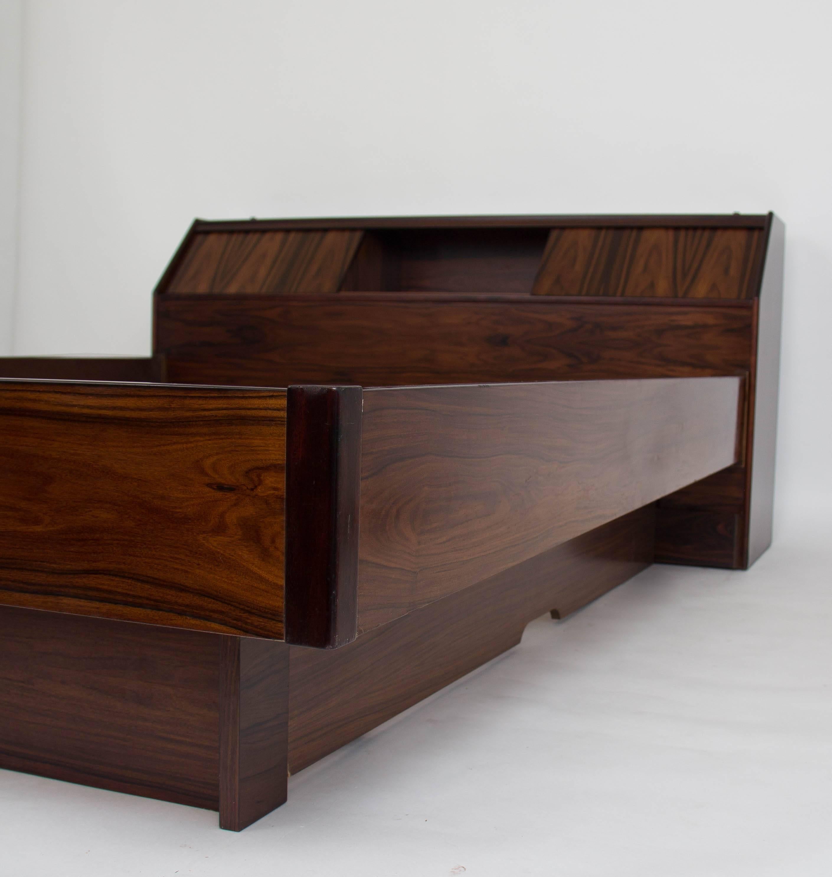 A queen-sized bed frame and headboard in highly figured rosewood, distributed and exported from Norway by Westnofa. The angled headboard has two sliding doors that open onto a storage compartment. The platform seems to float, with recessed siding in
