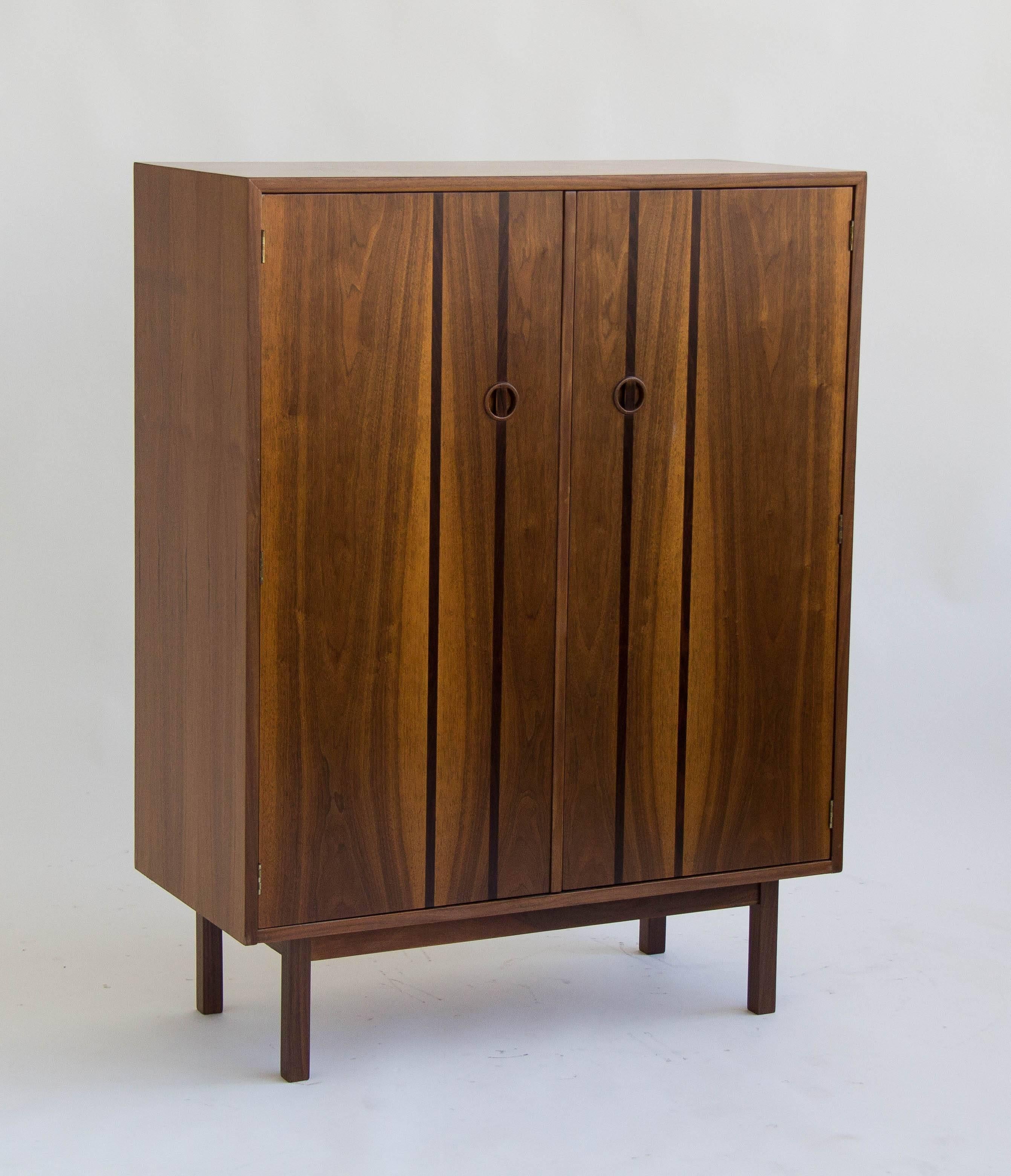 This highboy cabinet by H. Paul Browning for Stanley’s “Royal American” collection has a walnut case, resting on four recessed legs. The front has two doors with recessed round pulls and four slim bands of rosewood veneer detailing. Interior