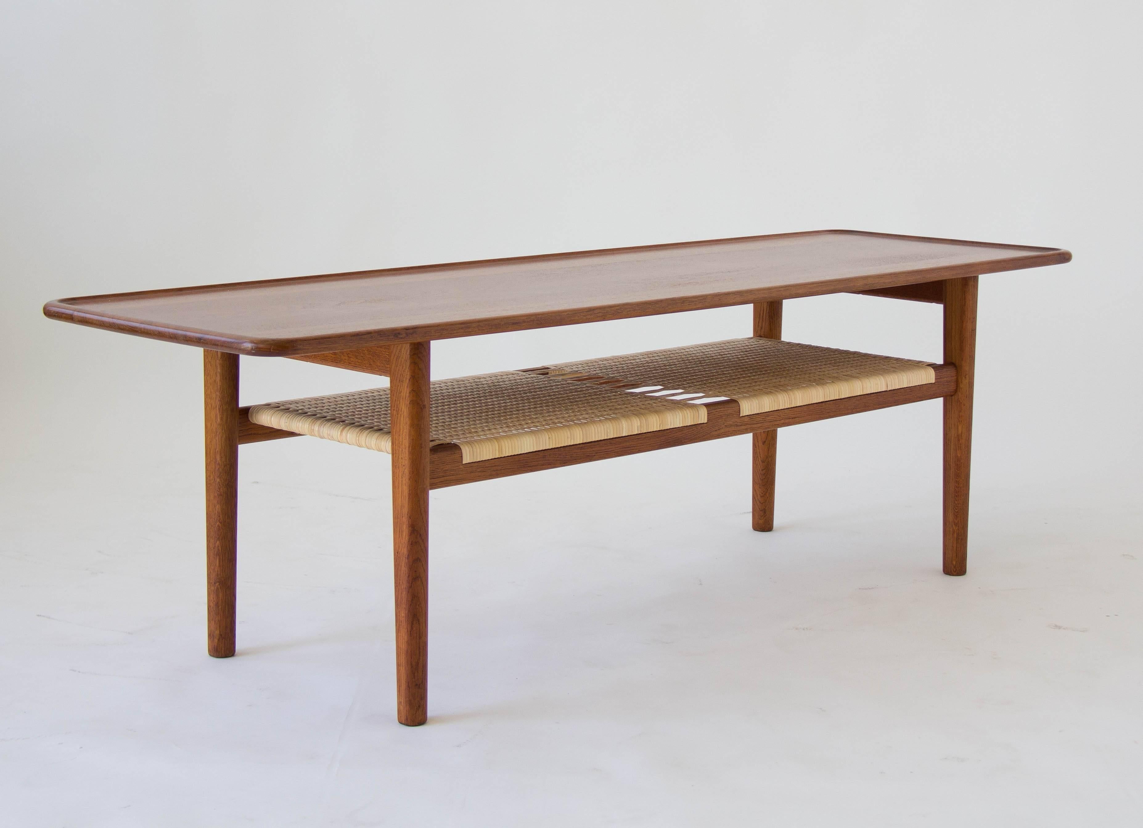 Gorgeous, unusually high coffee table designed by Hans Wegner for Andreas Tuck in 1955. A broad tabletop with a rounded and raised edge is crafted in solid teak and sits atop an oak base. The magazine shelf underneath is covered with woven cane.