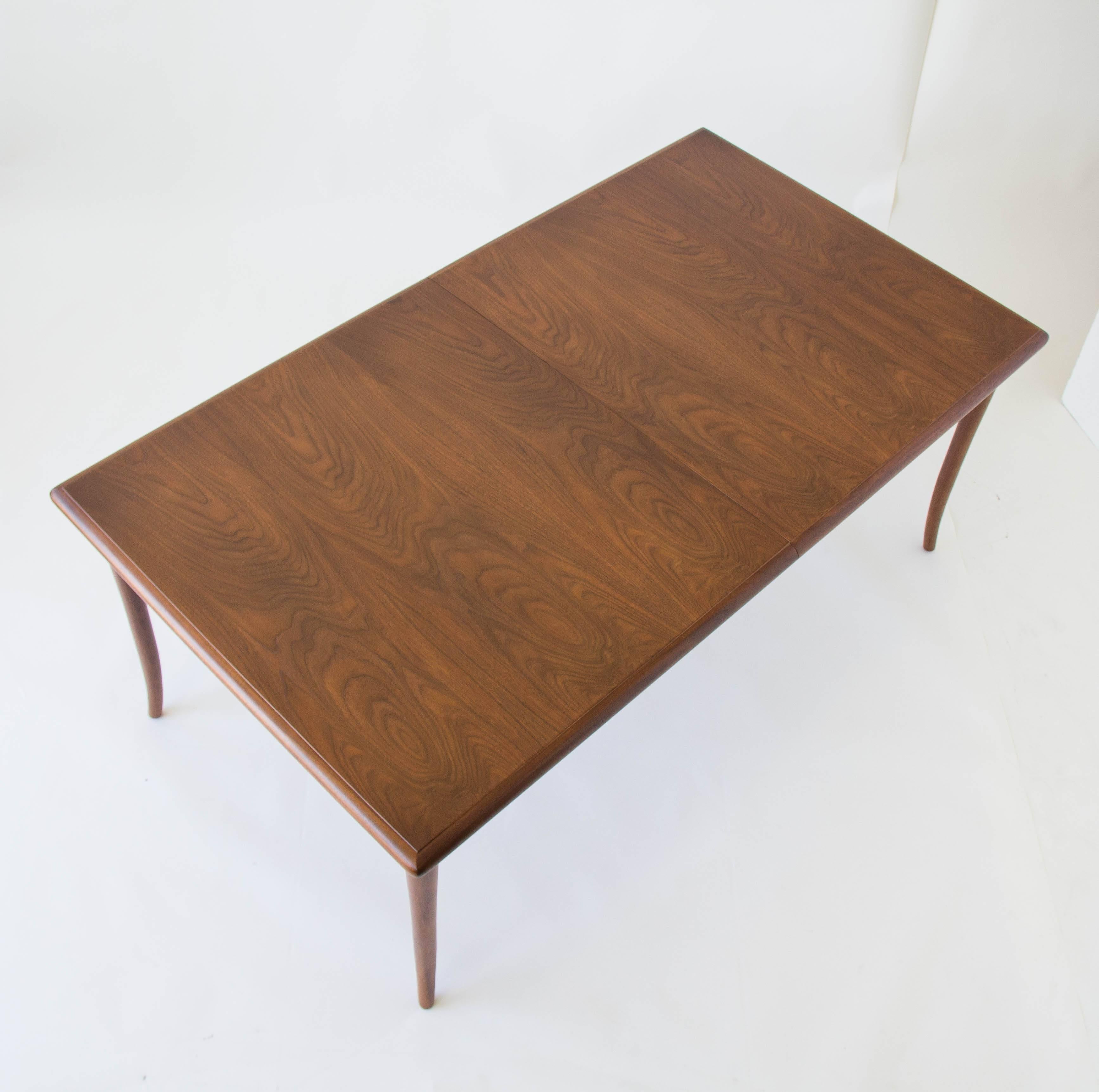 American made dining table by T. H. Robsjohn-Gibbings for Widdicomb showcasing beautiful walnut woodgrain. Deceptively simple design with a subtle raised tabletop, rounded edges and solid sabre legs that accommodates two insert leaves. Each walnut