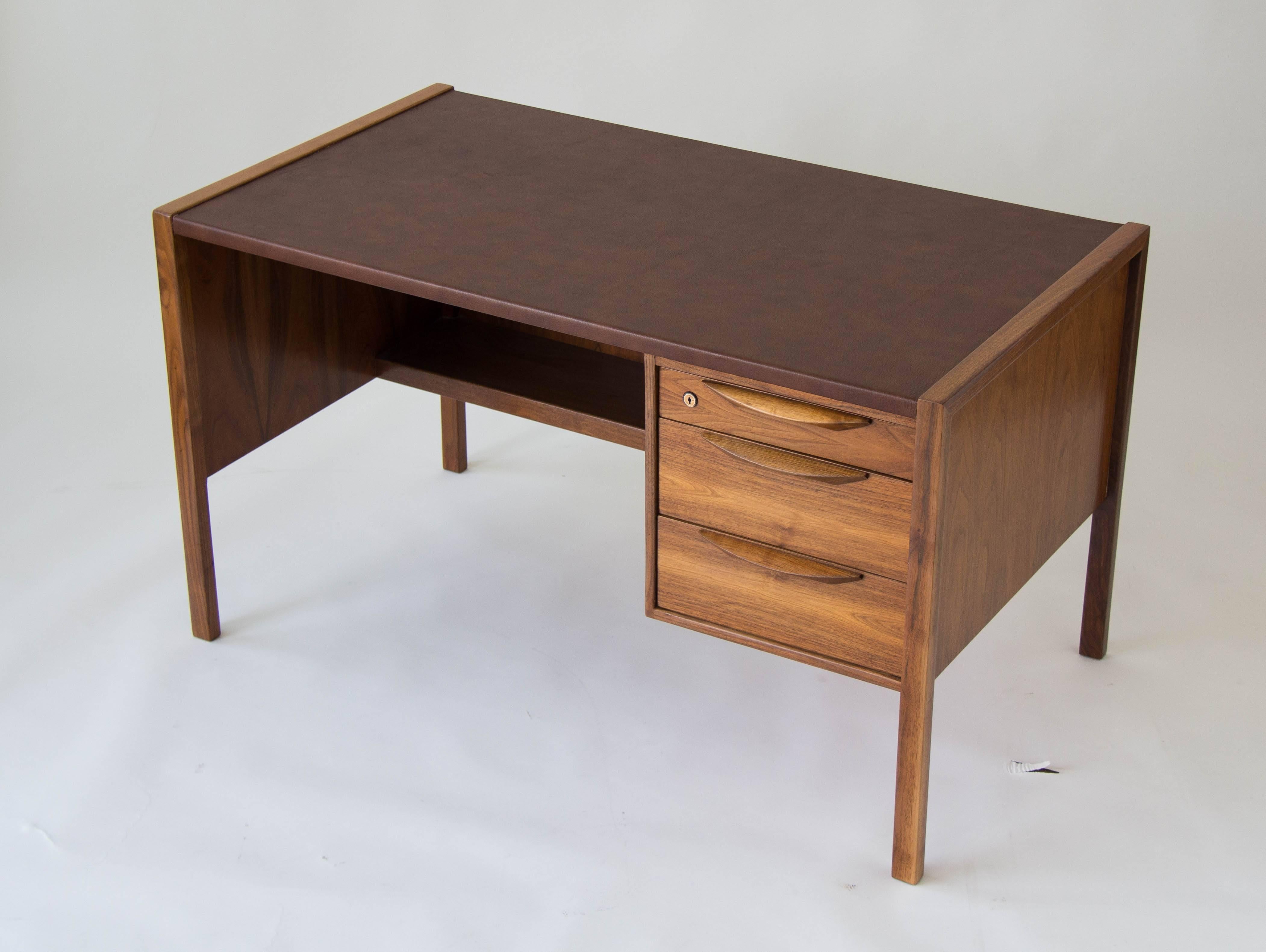 A modest walnut desk from Jens Risom with three drawers on the right-hand side, and a leather writing surface. Each drawer has a sculpted walnut pull and the top drawer locks for secure storage. Finished walnut panels cover the sides and back of the