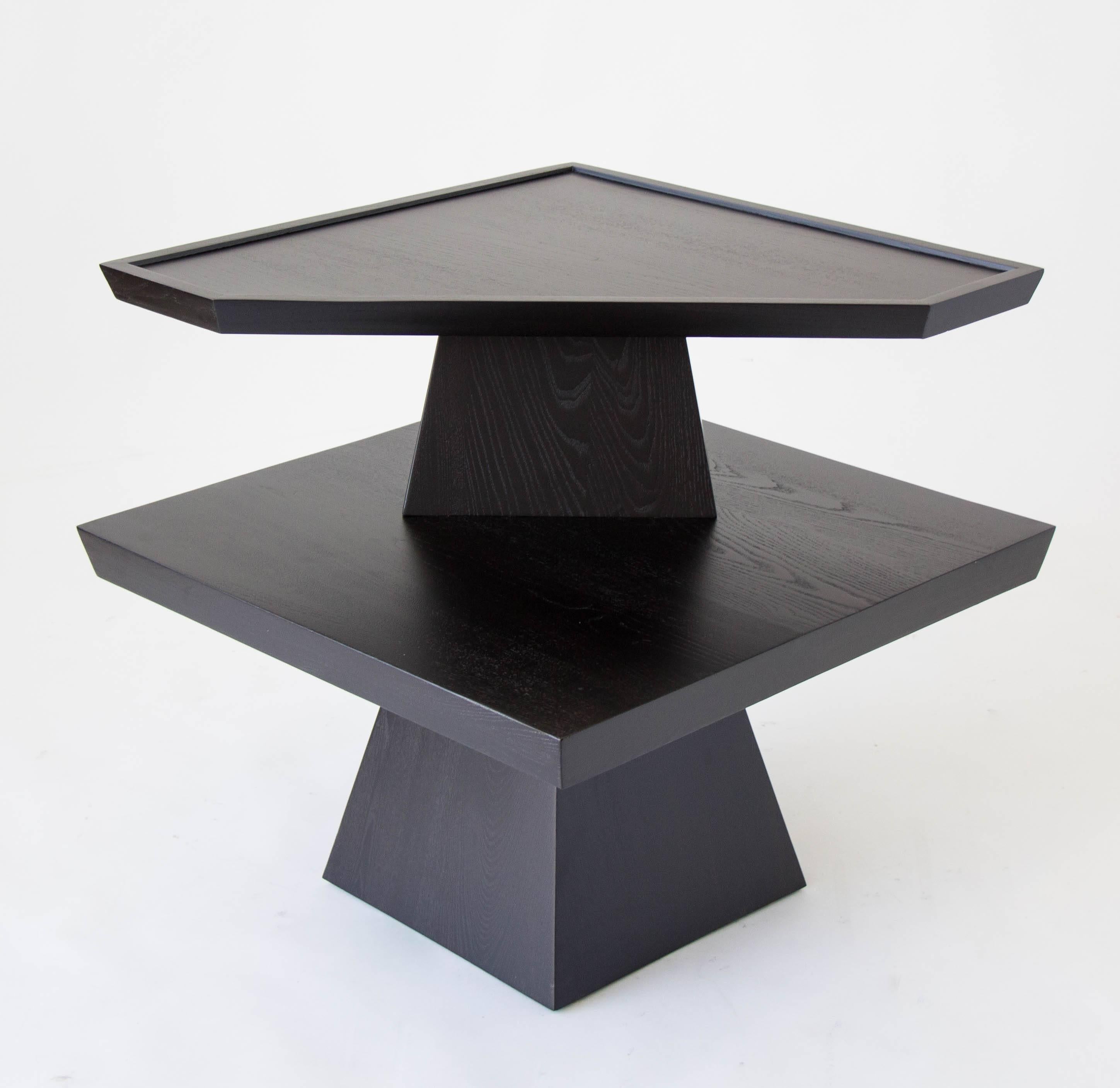 A two-tiered end or side table with irregular angles. The piece has a polygonal base with a wide tabletop, upon which is mounted a smaller pedestal and shelf with irregular sides. The smaller shelf has a raised lip. The entire piece has been