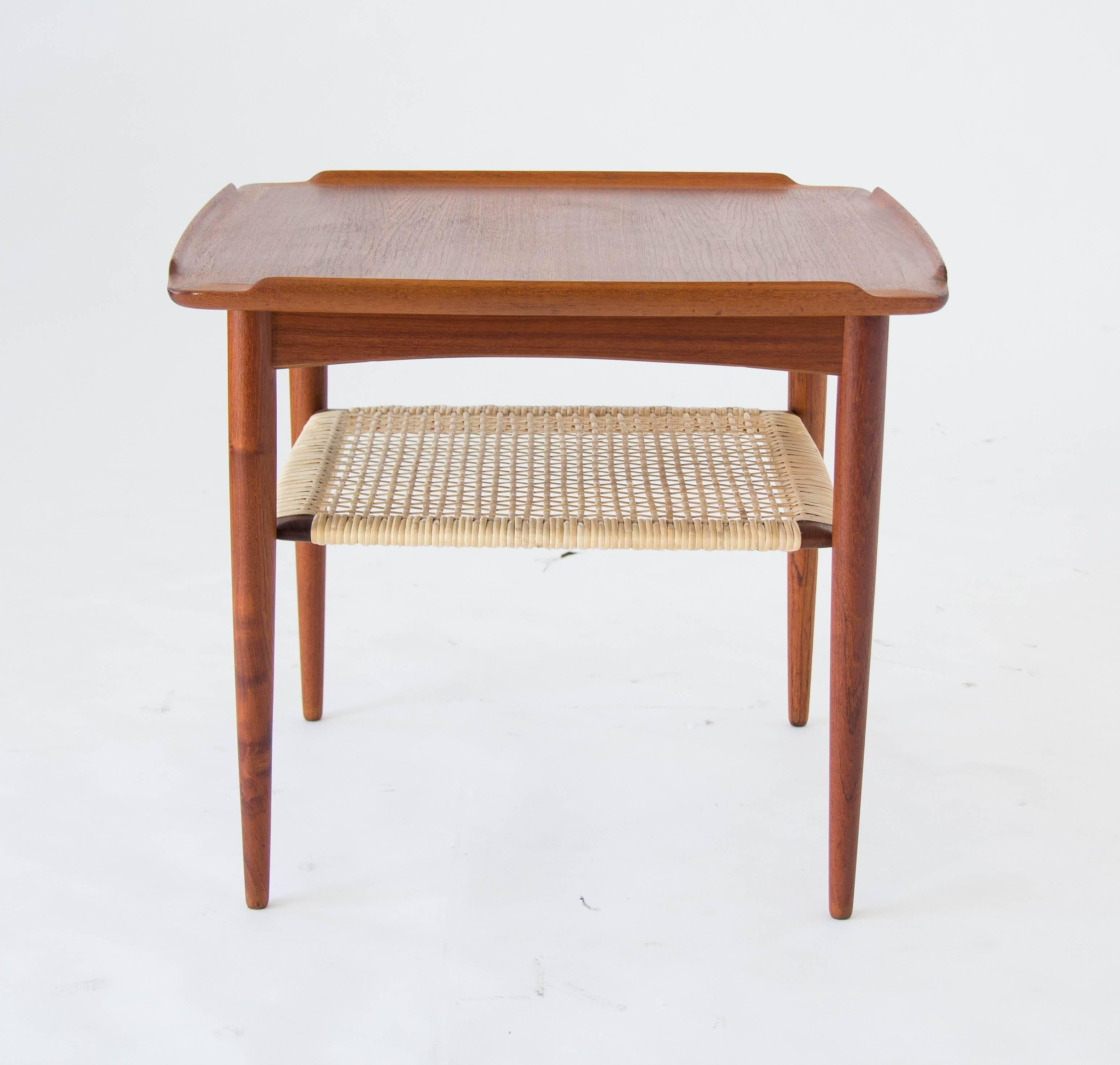 A square teak side table with a cane shelf. Designed by Poul Jensen and imported to the US by Selig, the table has a solid teak end piece with an upturned lip along each edge and an inlaid teak top. A shelf strung with woven cane is fixed to the