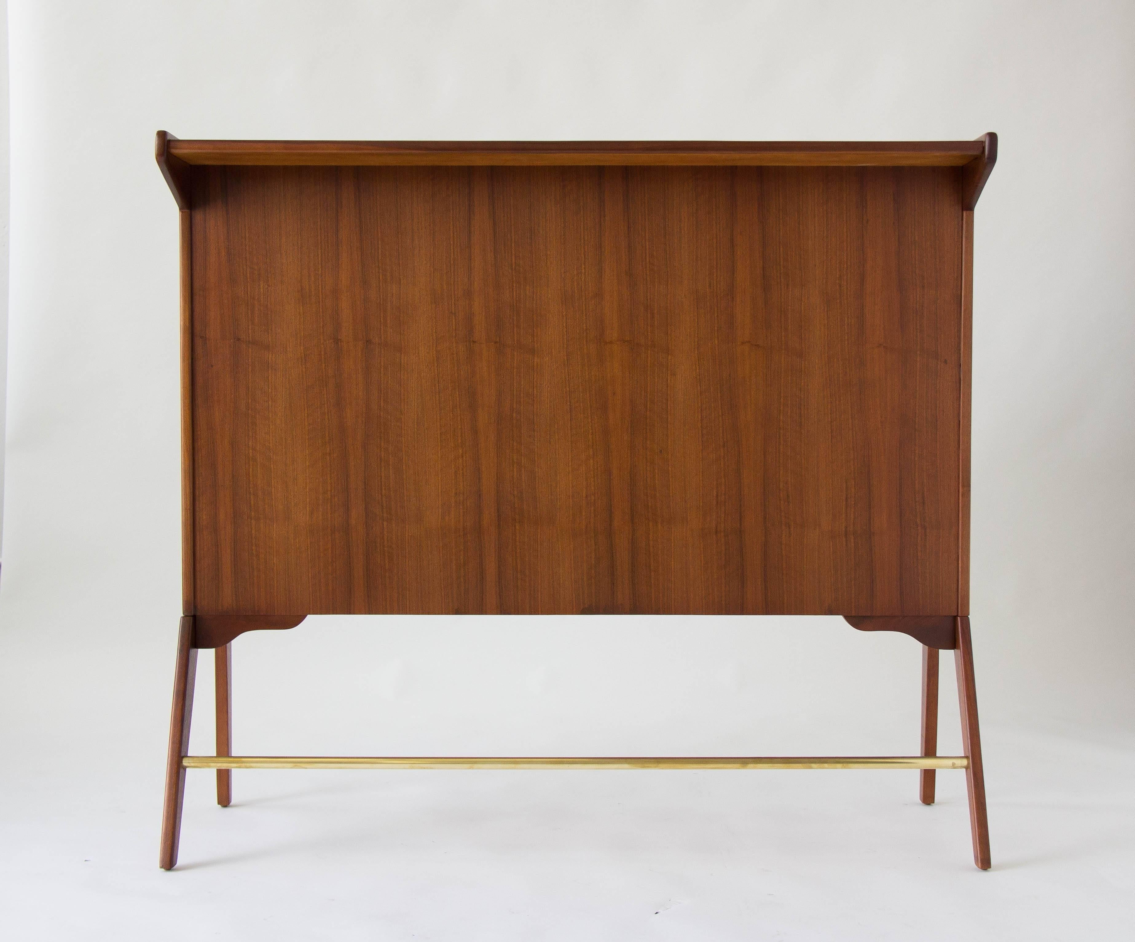 A high walnut serving bar with stylistically kicked-out front legs. The bar top has an overhang on the front-facing side and is open on the other side with a workspace and two storage compartments. A brass stretcher decorates the front of the piece