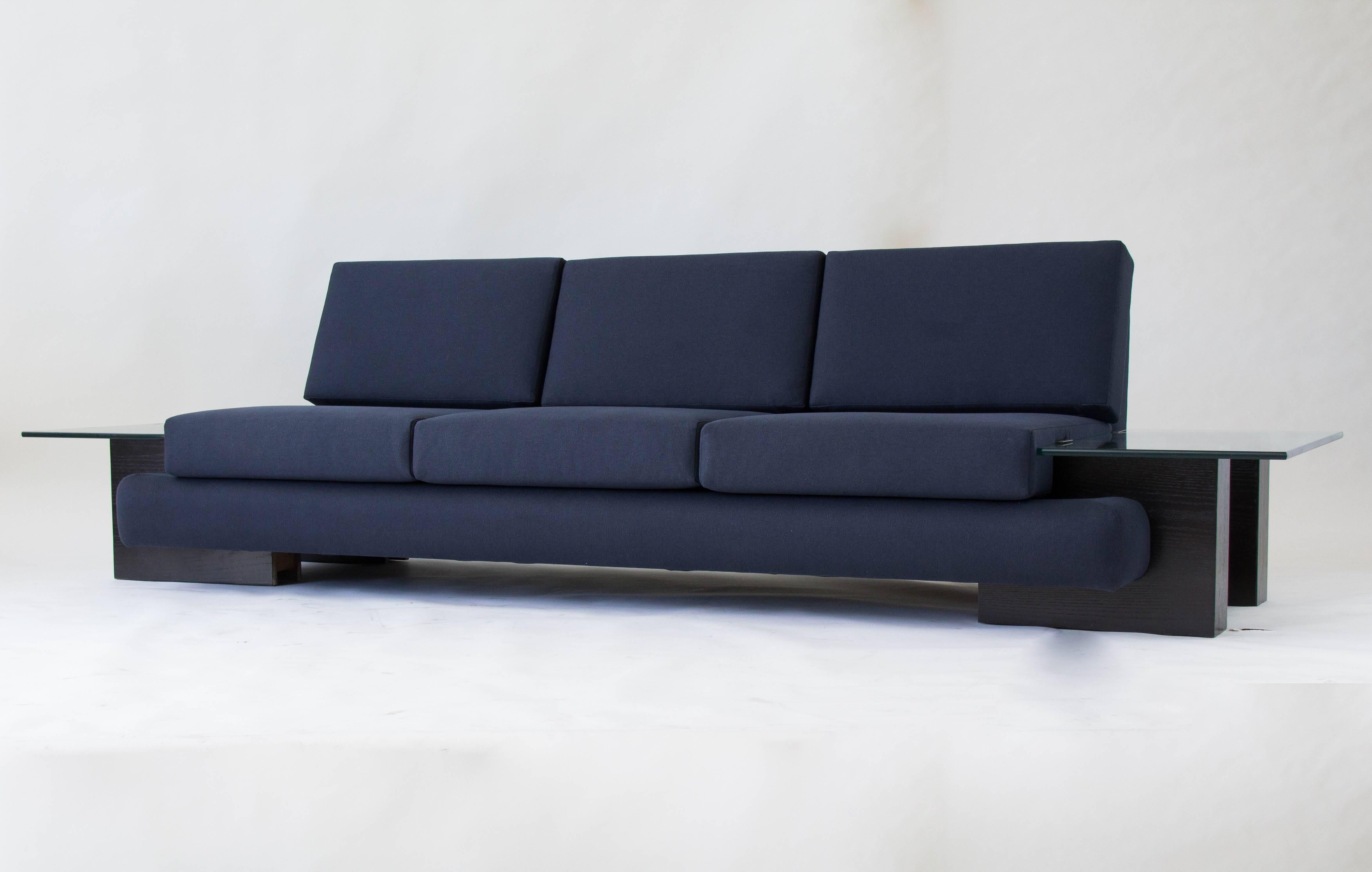 From midwestern manufacturer Kroehler, this sofa from 1978 has a rounded, upholstered frame in Oxford blue cotton. The platform sits on four molded wood blocks with an ebonized finish, and a built-in end table of thick-cut glass is mounted on either