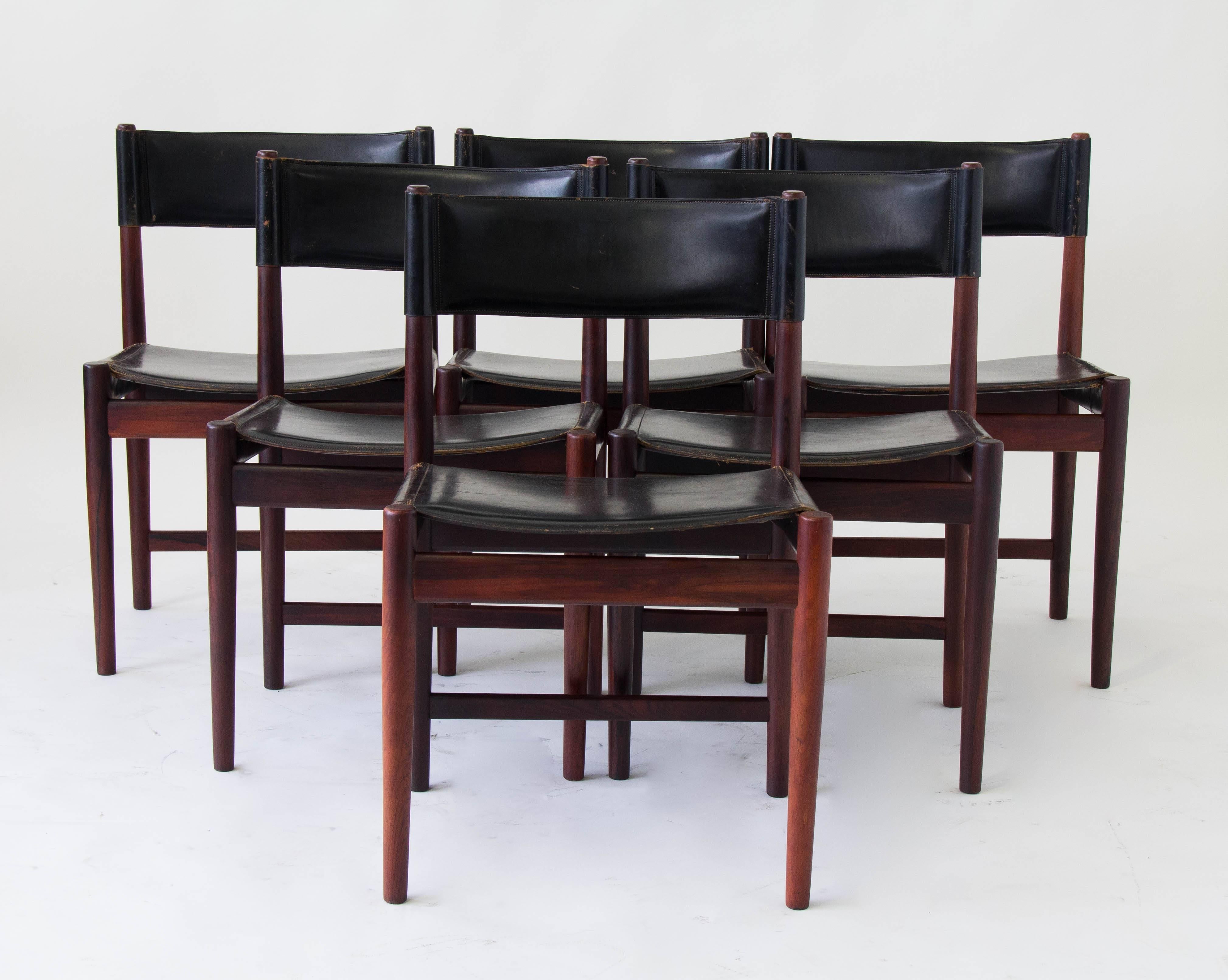 Six rosewood dining chairs designed by Kurt Østervig and produced by Sibast furniture. Each chair has a straight back and tapered dowel legs. The frame is wrapped in black saddle leather to form the backrest and seat cushion. Sibast manufacturer’s