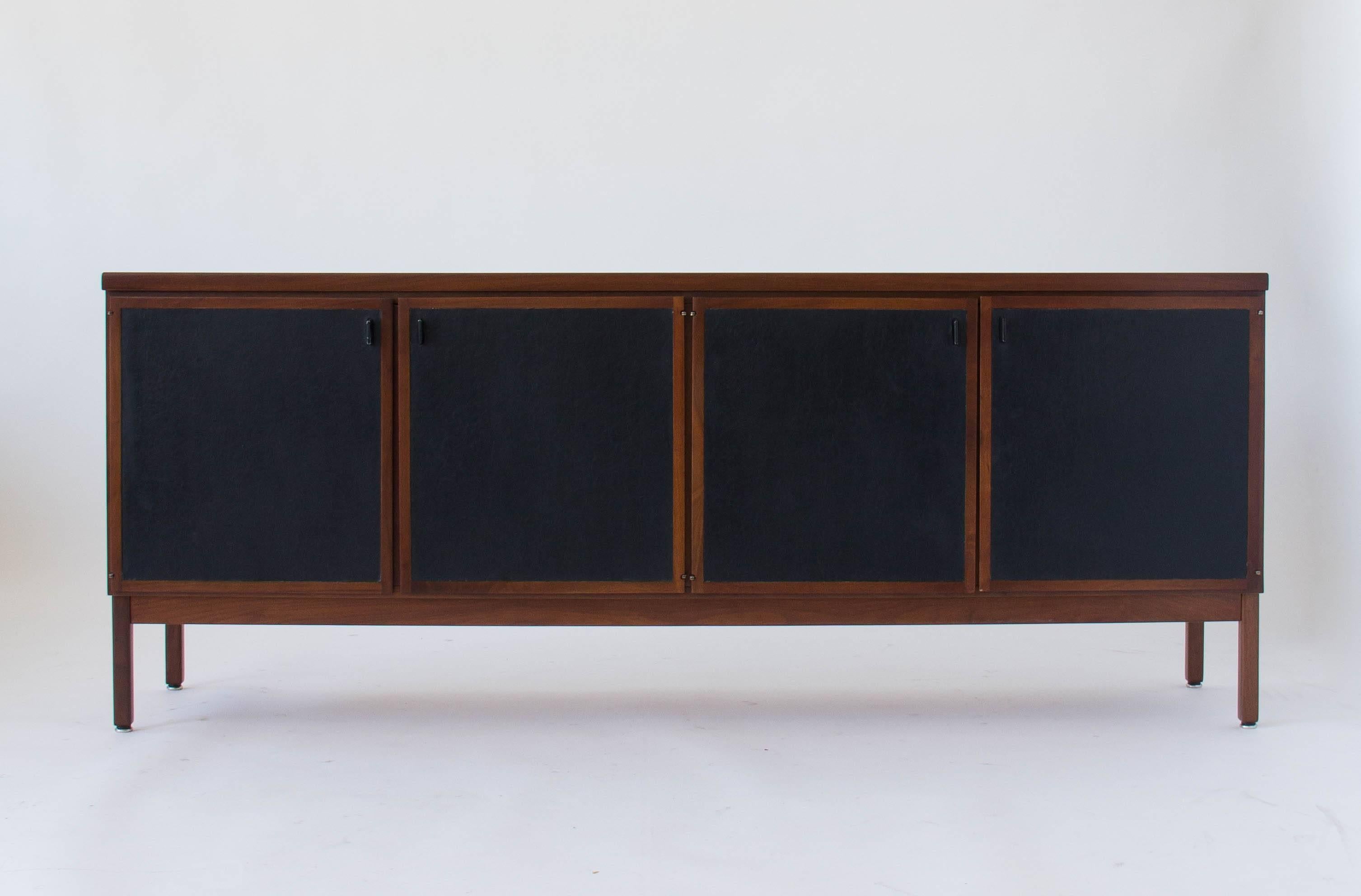 A Mid-Century modern credenza in walnut wood, standing on four square legs of solid walnut. Four leather-paneled doors with flat pulls open on two storage bays. The left-hand bay has two shallow drawers and an open storage space inside; while the