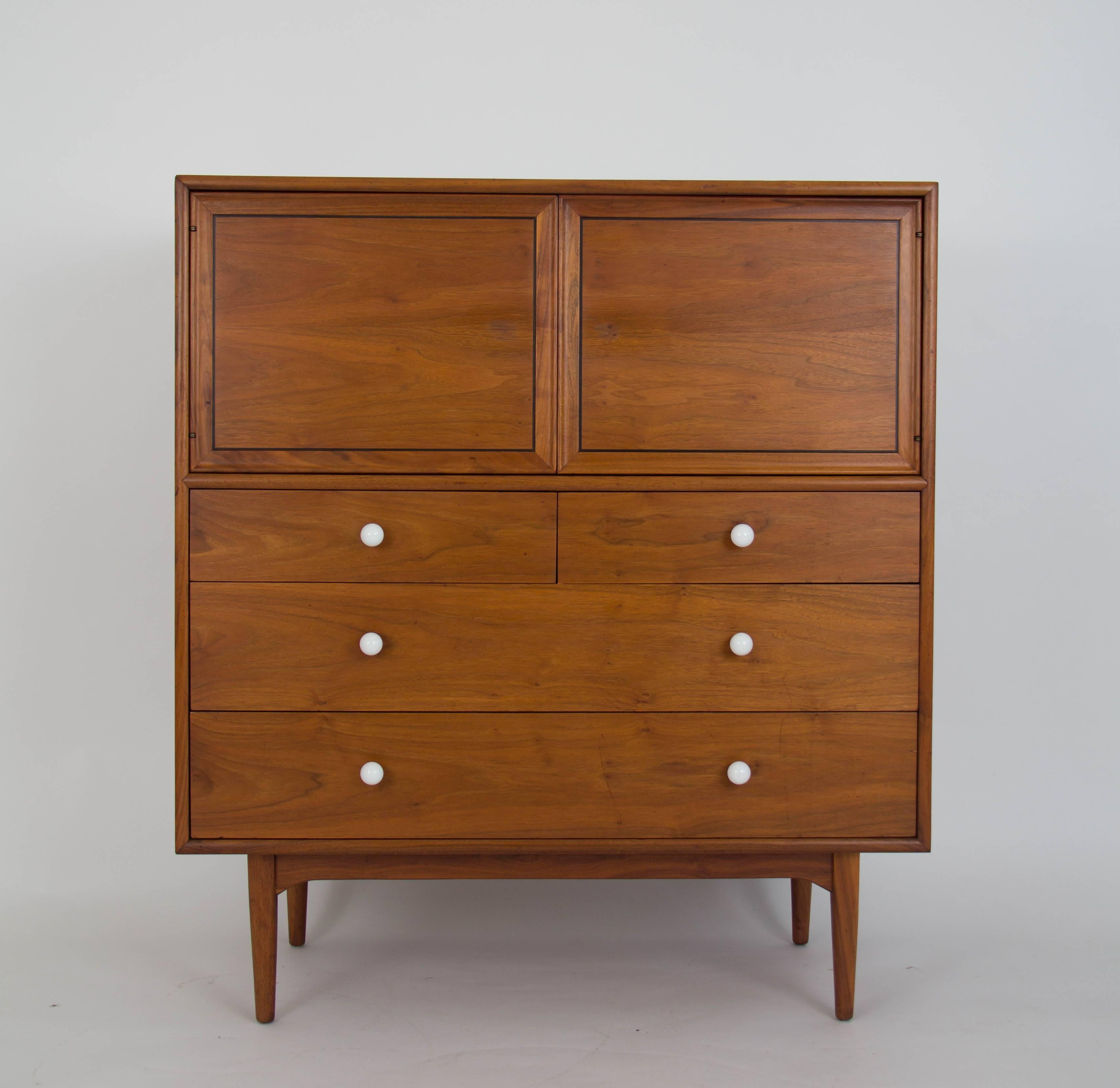 A tall, walnut highboy dresser and vanity from Drexel’s popular Declaration collection designed by Kipp Stewart and Stewart MacDougall. This example has a walnut case holding five subdivided drawers with original white ceramic knobs on brass spacers