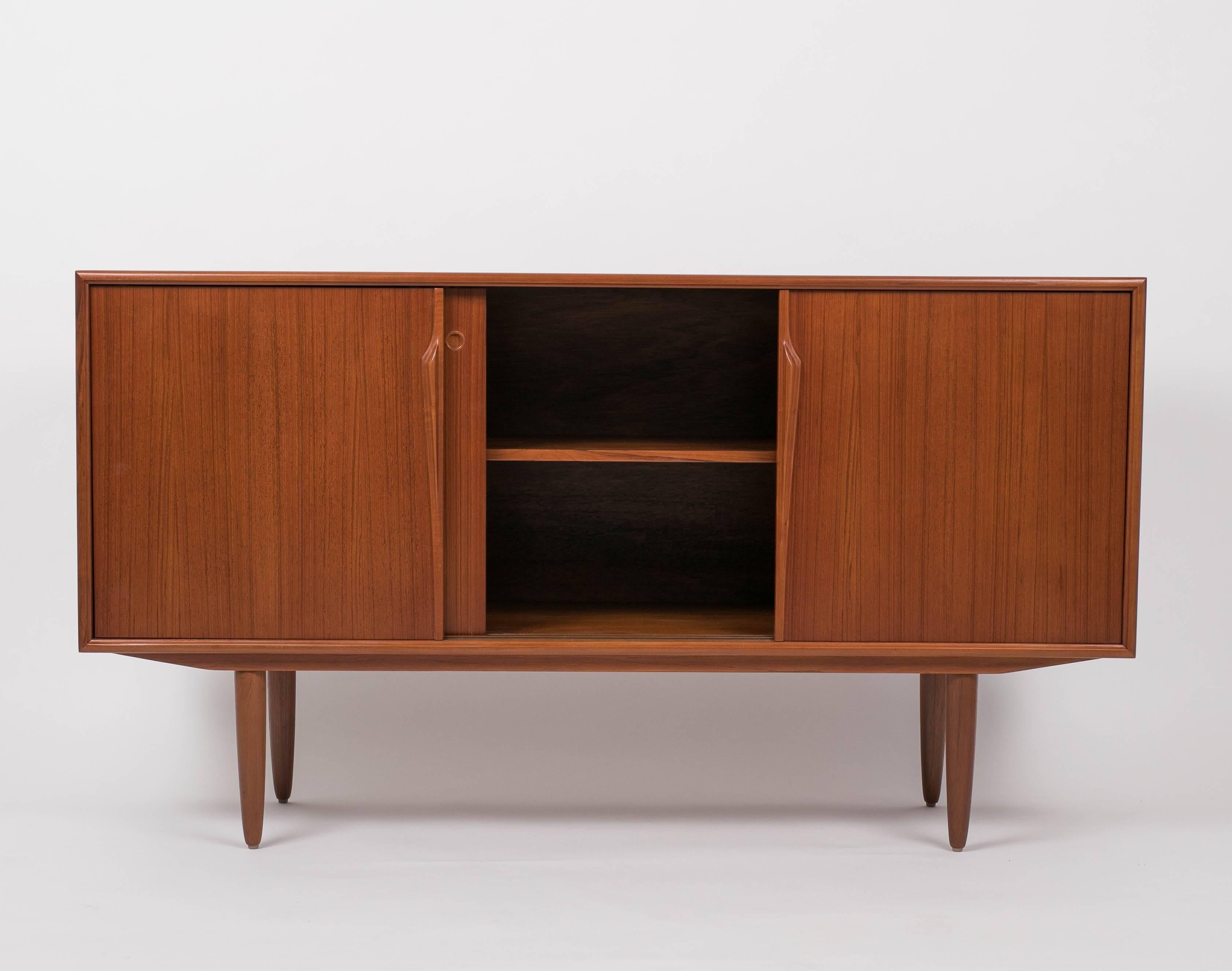 A Gunni Omann-designed credenza in teak wood. The compact piece has unusually high solid teak legs with a distinctive bullet shape and three sliding panels across its face. The outer doors have ridged pulls and slide freely along the length of the