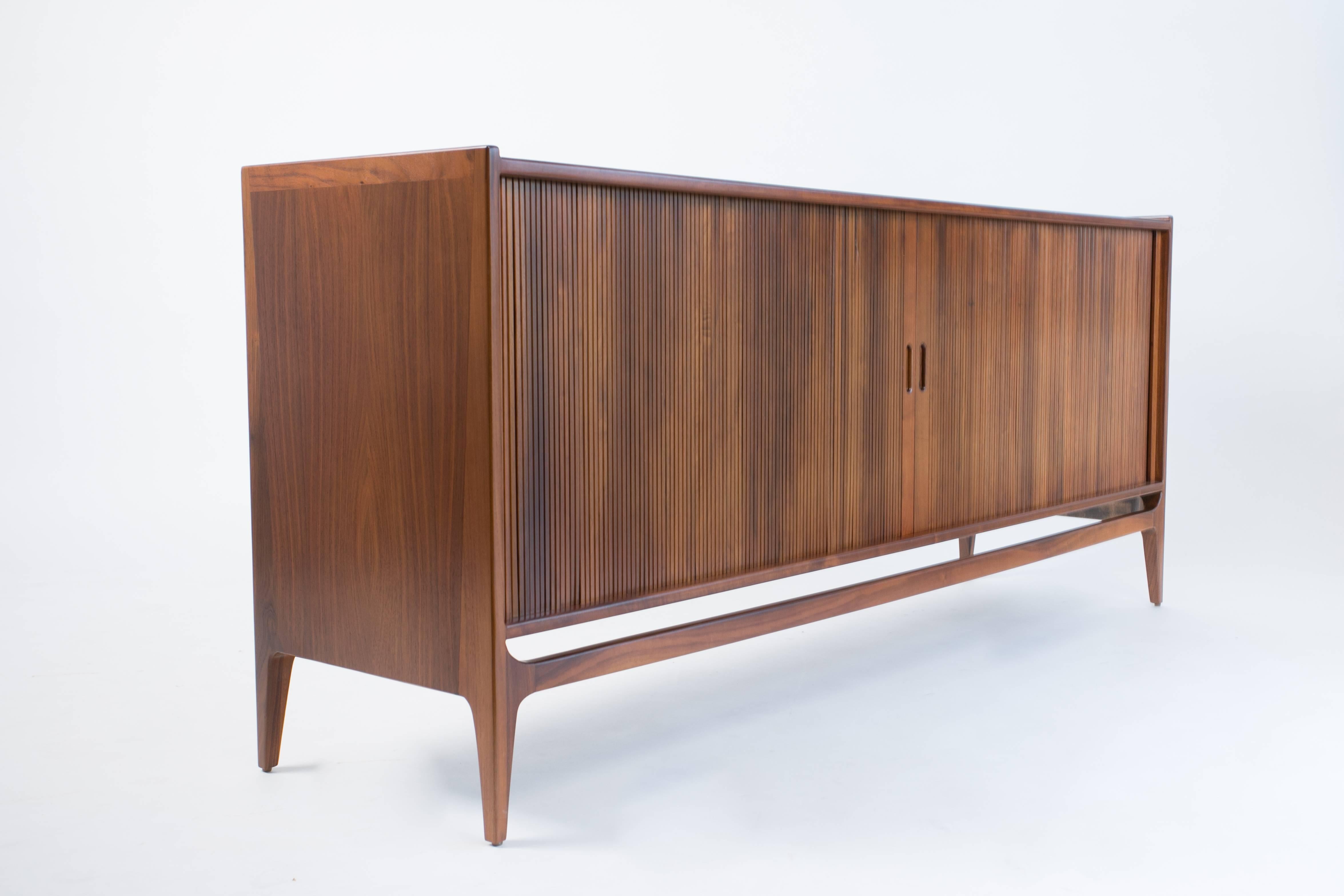 A dark walnut credenza by Richard Thompson for Glenn of California with tambour doors and modular interior shelving. The tapered legs of the piece sit flush with the walnut case, and the tambour doors with recessed pulls open onto three storage