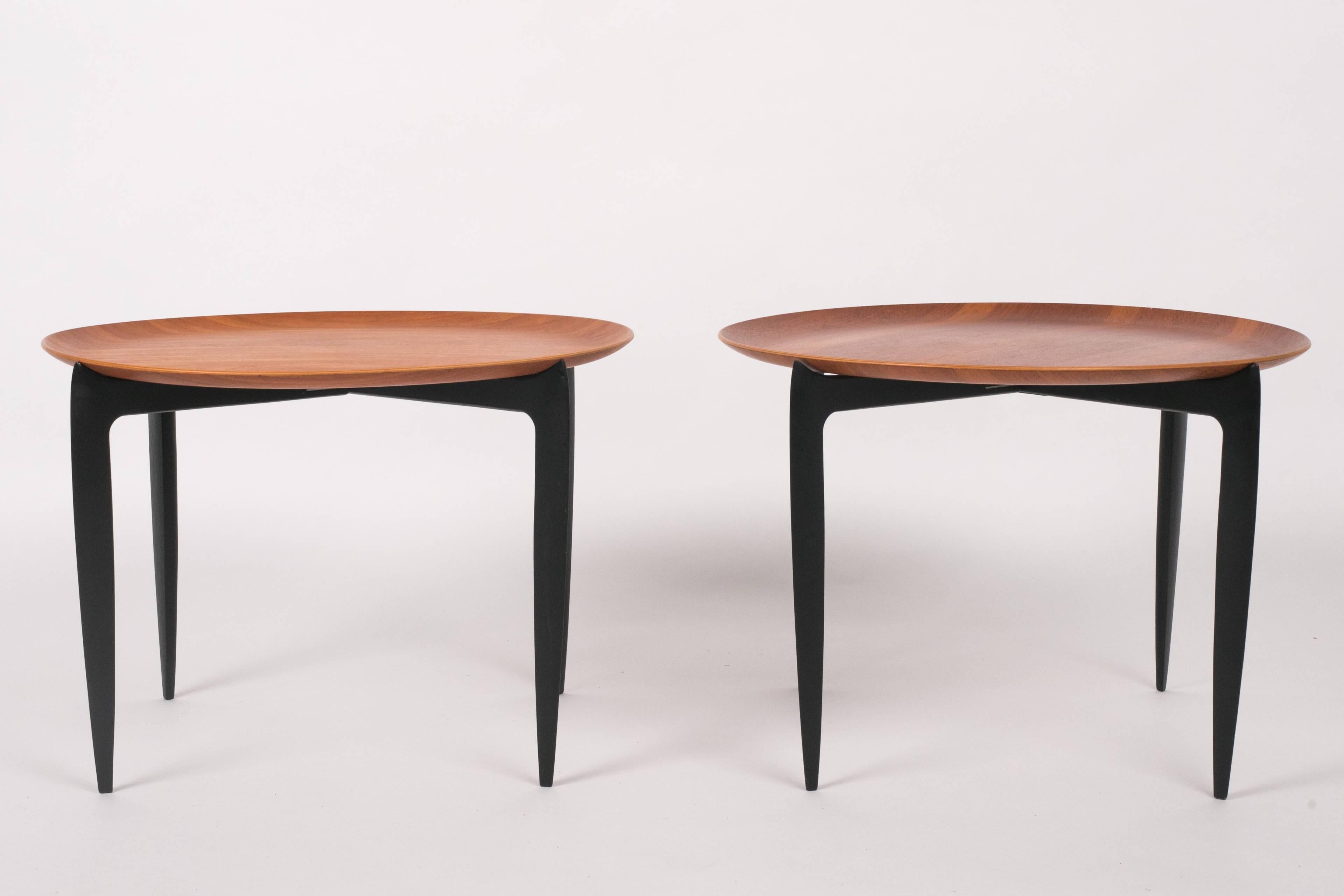 An in-house design for Fritz Hansen, by Svend Åage Willumsen and H. Engholm, this pair of teak tray tables has ebonized, folding bases. Each table has a wide round top of molded teak, with a slightly raised lip. The folding base has four tapered