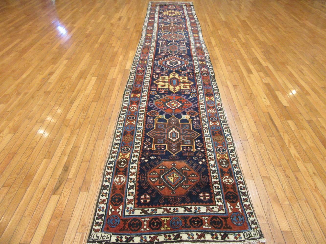 This is a long genuine hand-knotted antique Persian Heriz runner rug. It has an inscription on the corners of one end indicating it was made in 1923. The rug has a simple geometric pattern on a navy blue background and rusty red border. It measures