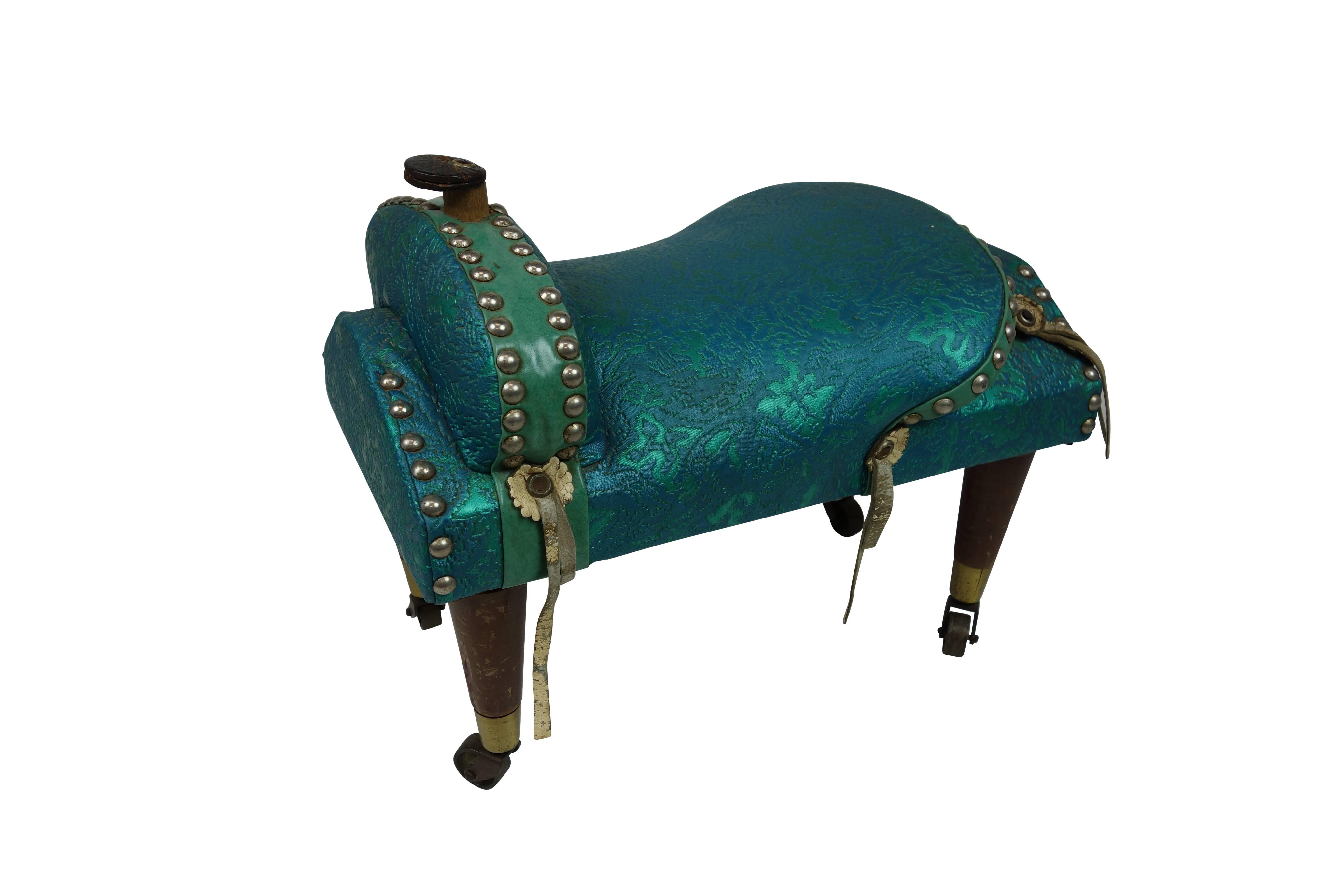 This is a unique teal upholstered and riveted saddle foot stool on casters.