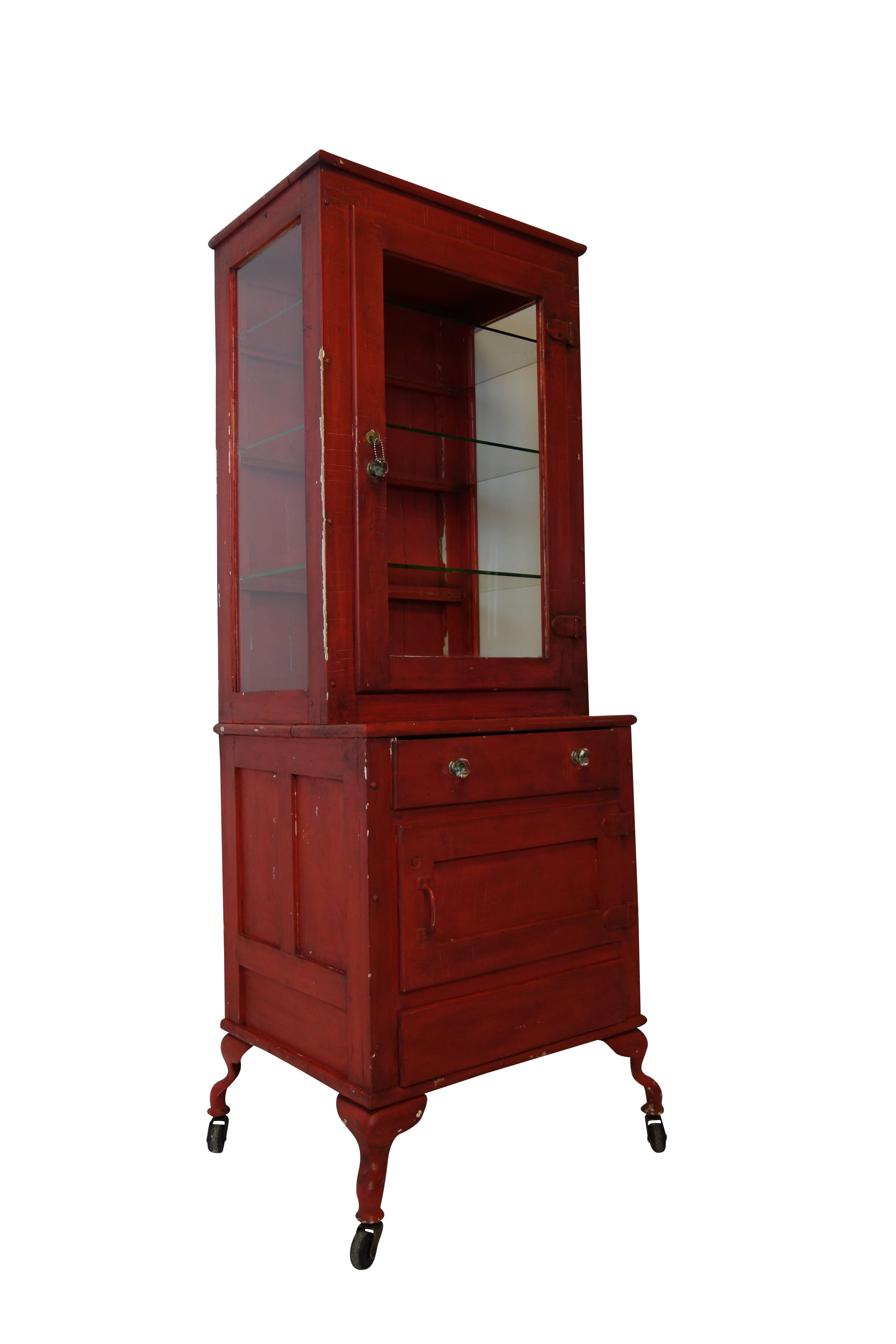 This is a unique red painted medical cabinet with cabriole legs on casters. The set back three-sided glass display has the original lock and key. The lower portion has a hinged cabinet space for storage and two drawers. The bottom drawer appears to