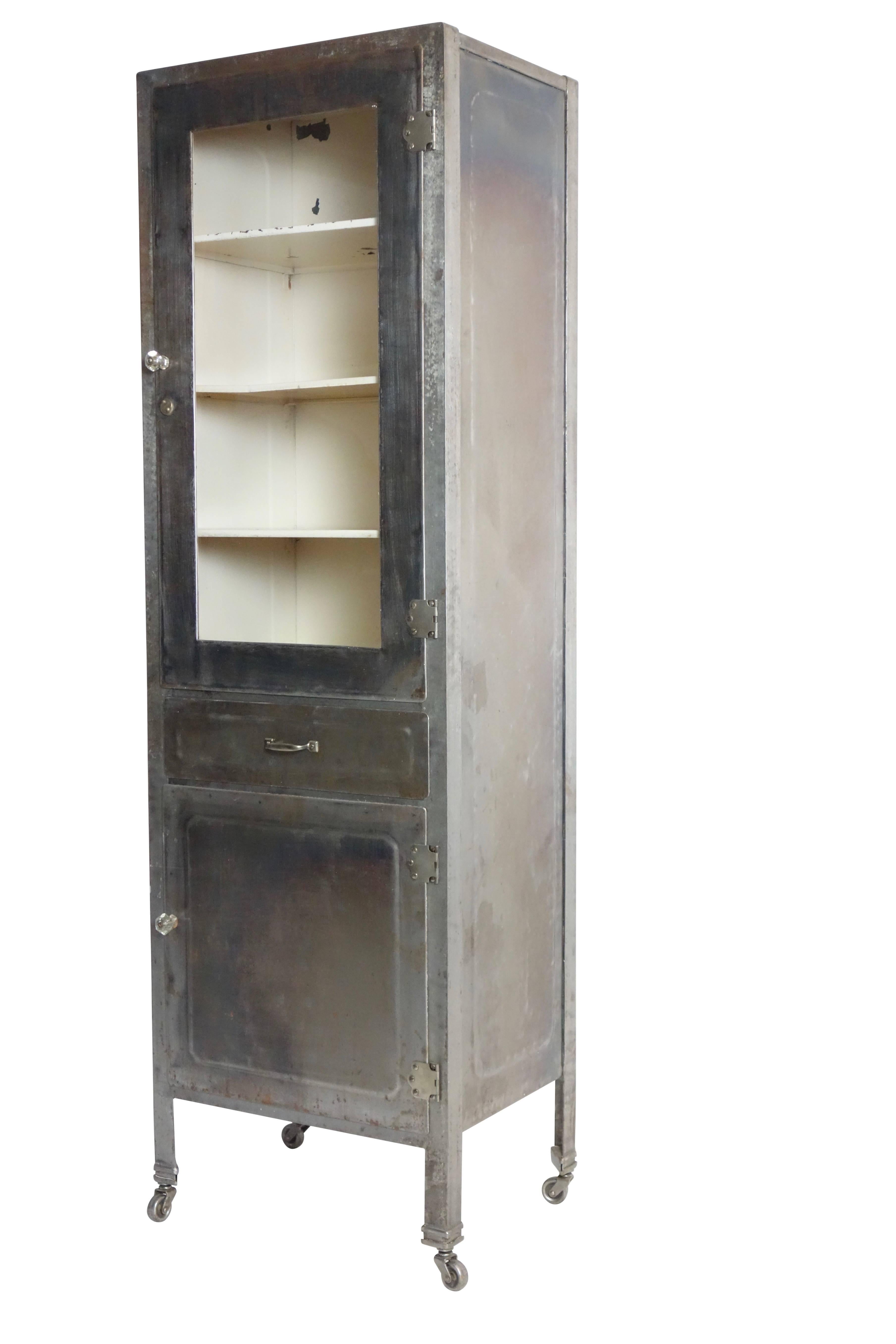This is a steel medical cabinet on casters with three interior shelves behind a glass door. The lower compartment has two interior shelves.