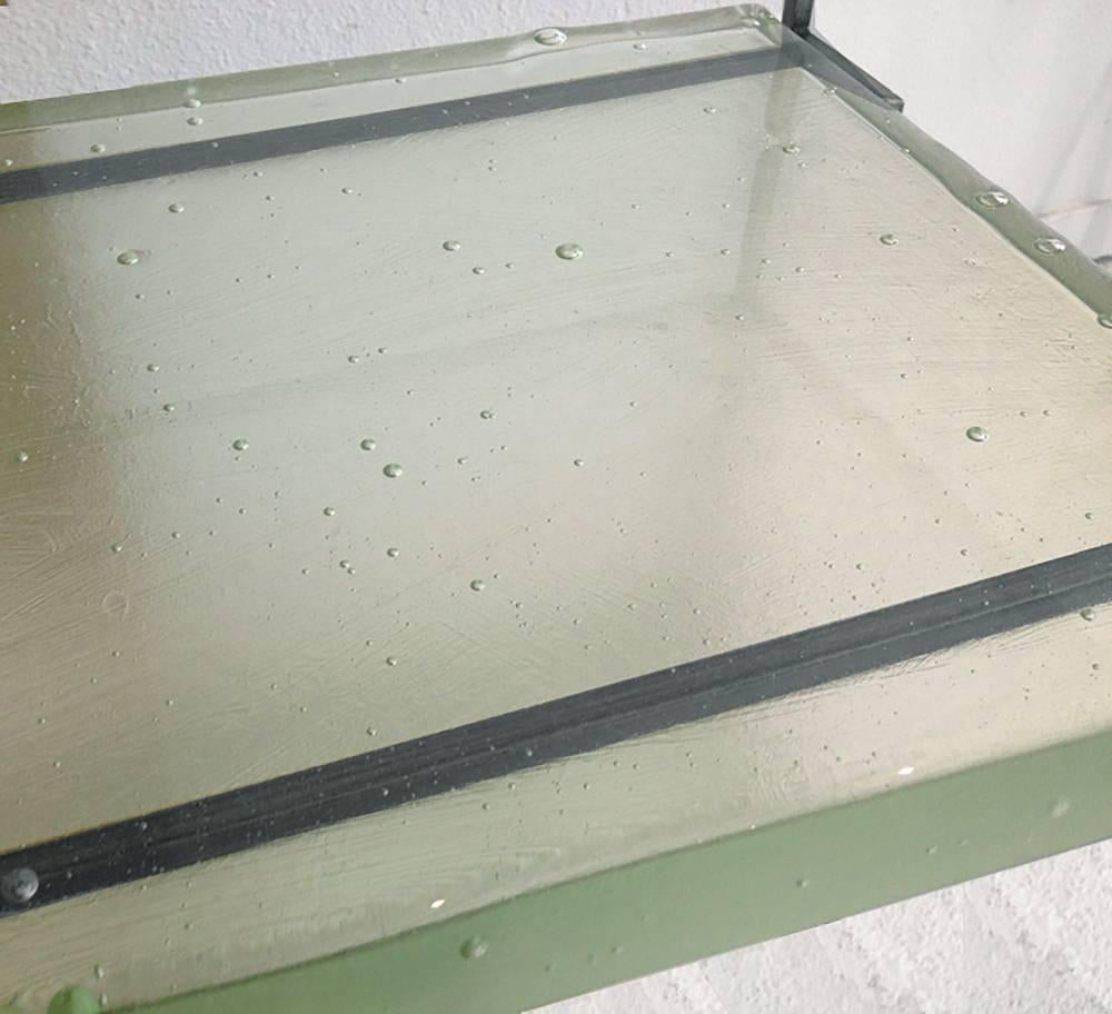 Light green hand-cast glass slab floating in a blackened steel frame with a glass top. The hand-cast glass has natural bubbles and imperfections.