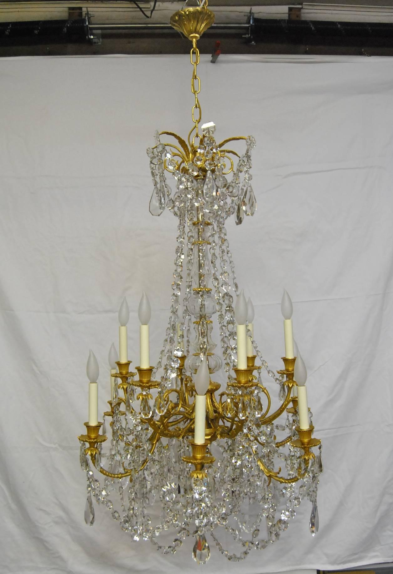 A gorgeous large scale French Empire crystal chandelier. The body of this chandelier is done in gilded bronze and has two layers of arms for a total of 12 arms. Arms are scrolled and hold various size crystals, some shaped. The center of the