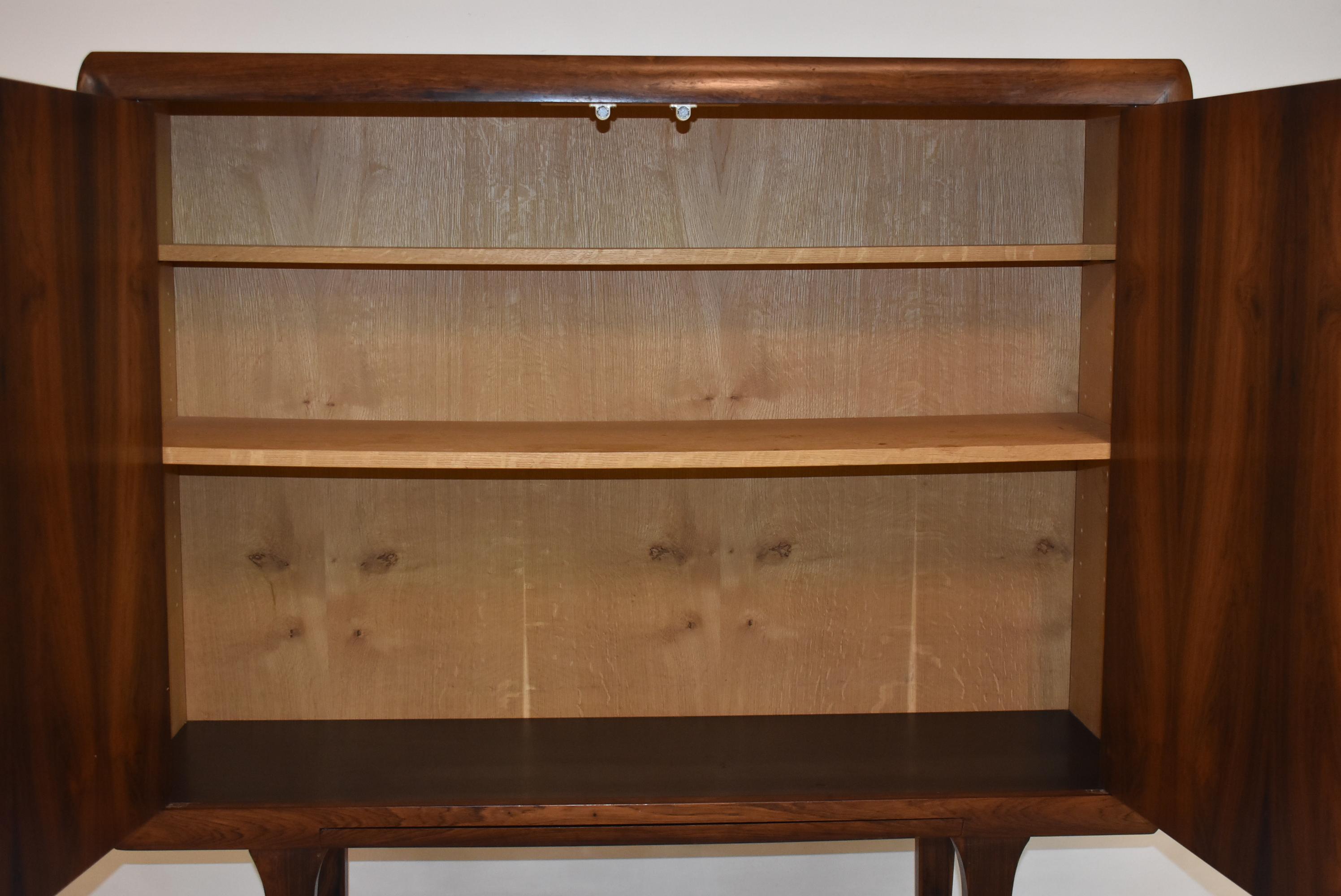 2-door bar or liquor cabinet by Ib Kofod Larsen constructed of figured rosewood. The cabinet sits atop contoured legs with cross stretcher supports, and features carved wood door pulls, 2 adjustable shelves, a black laminate interior bottom and a
