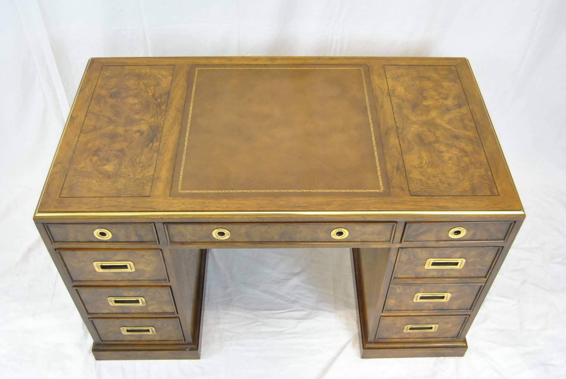 A Classic Campaign style desk by Drexel. The desk is done in burled wood with a leather insert on top and brass trim at top edge. There are three shallow drawers along the top. Along the left side there are three drawers and along the right side