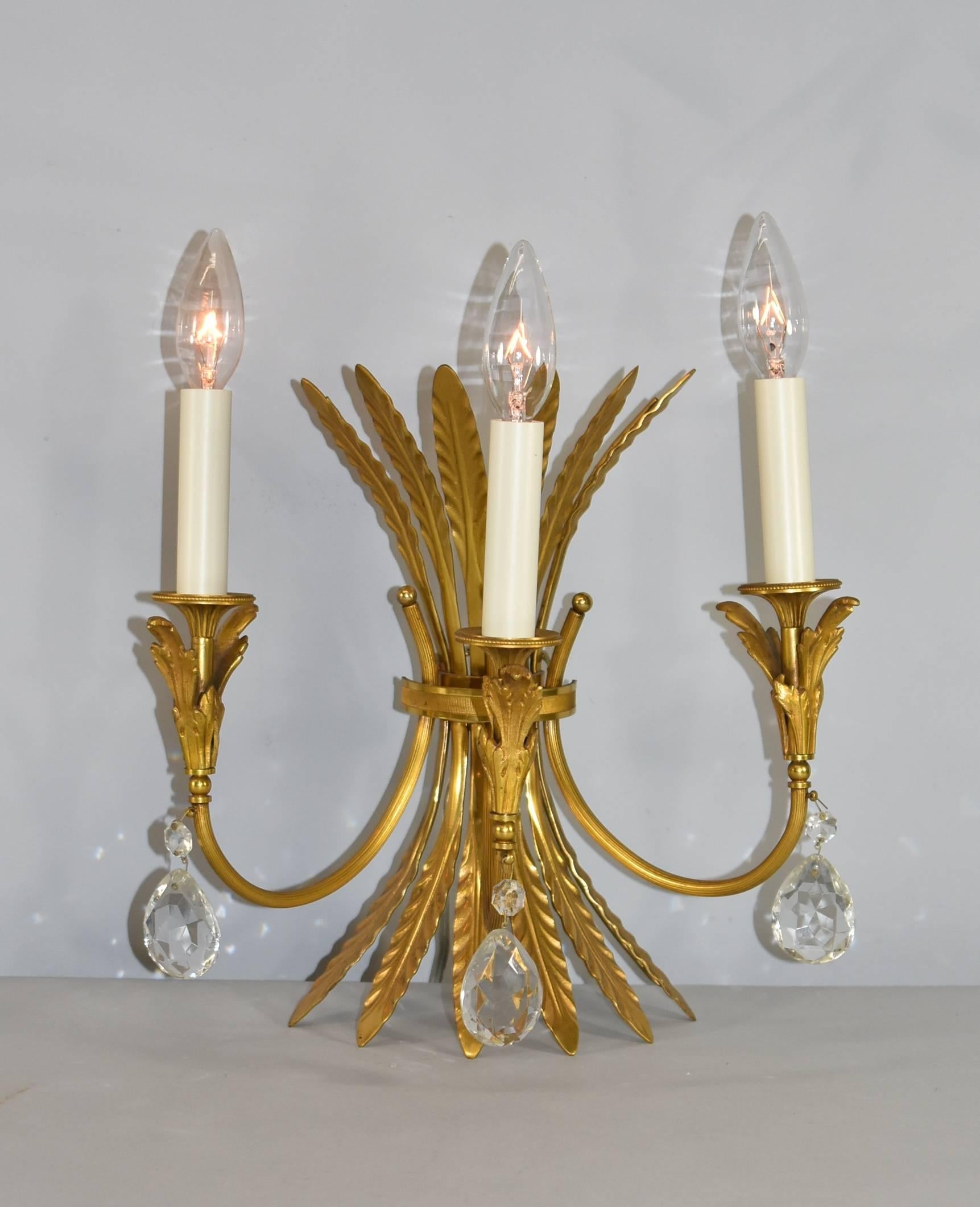 A beautiful pair of Italian neoclassic style three-arm brass wall sconces with cut crystal teardrop accenting. These sconces feature a sheaf body with leaf accents below the brass bobeches holding the cream candlesticks. You can add shades for a