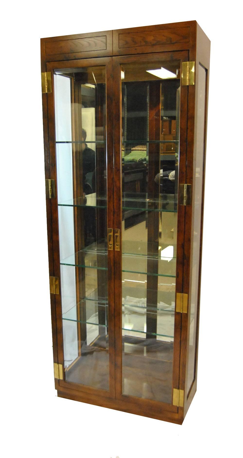 A great pair of Campaign style curio display cabinets. The cabinets feature two beveled glass doors which open to an interior with a mirrored back. There are 4 thick glass shelfs in each and two lights in the top. Heavy brass hardware adorns the