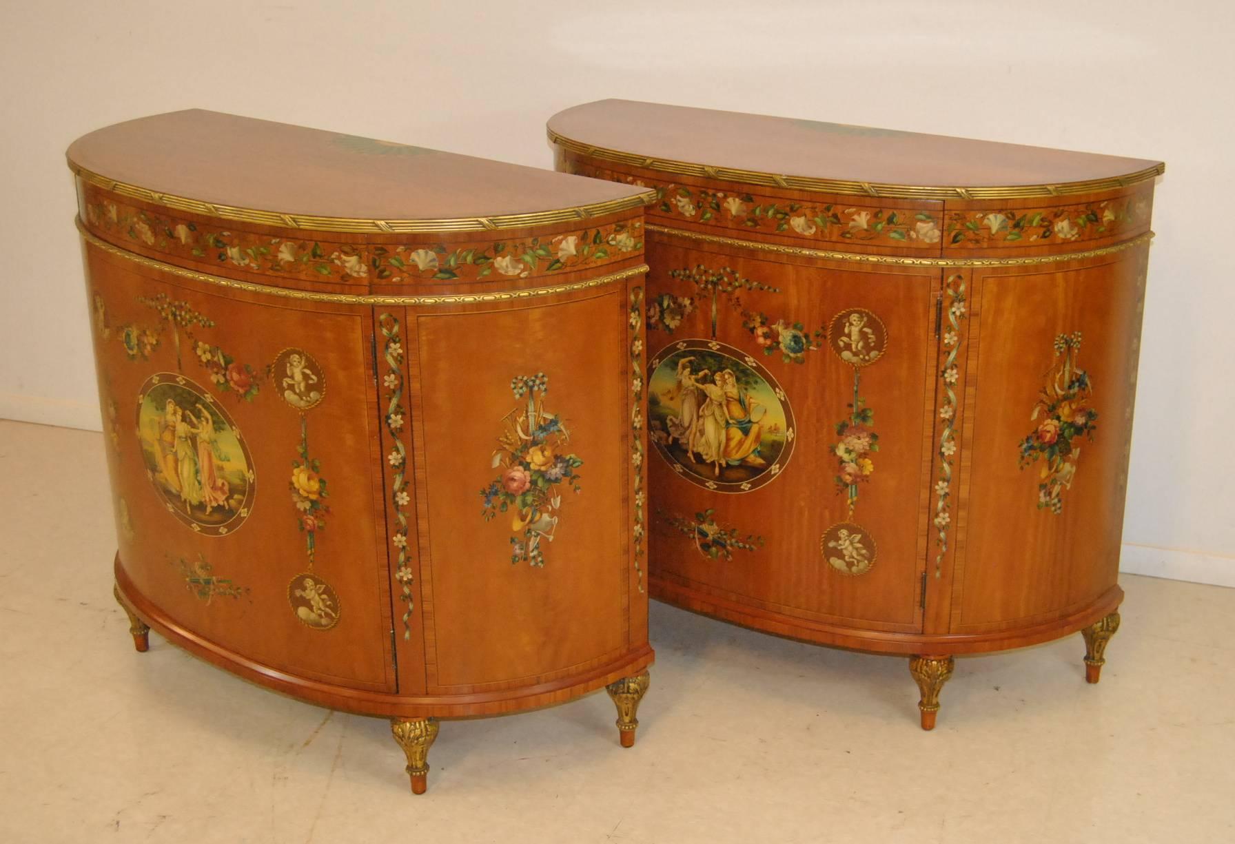 A gorgeous pair of Satinwood French Adam style commodes by Irwin Furniture, circa 1937. Sure to add style and elegance to any room in your home. These stunning commodes feature delicately hand-painted winged cherubs and fair maidens, one drawer that