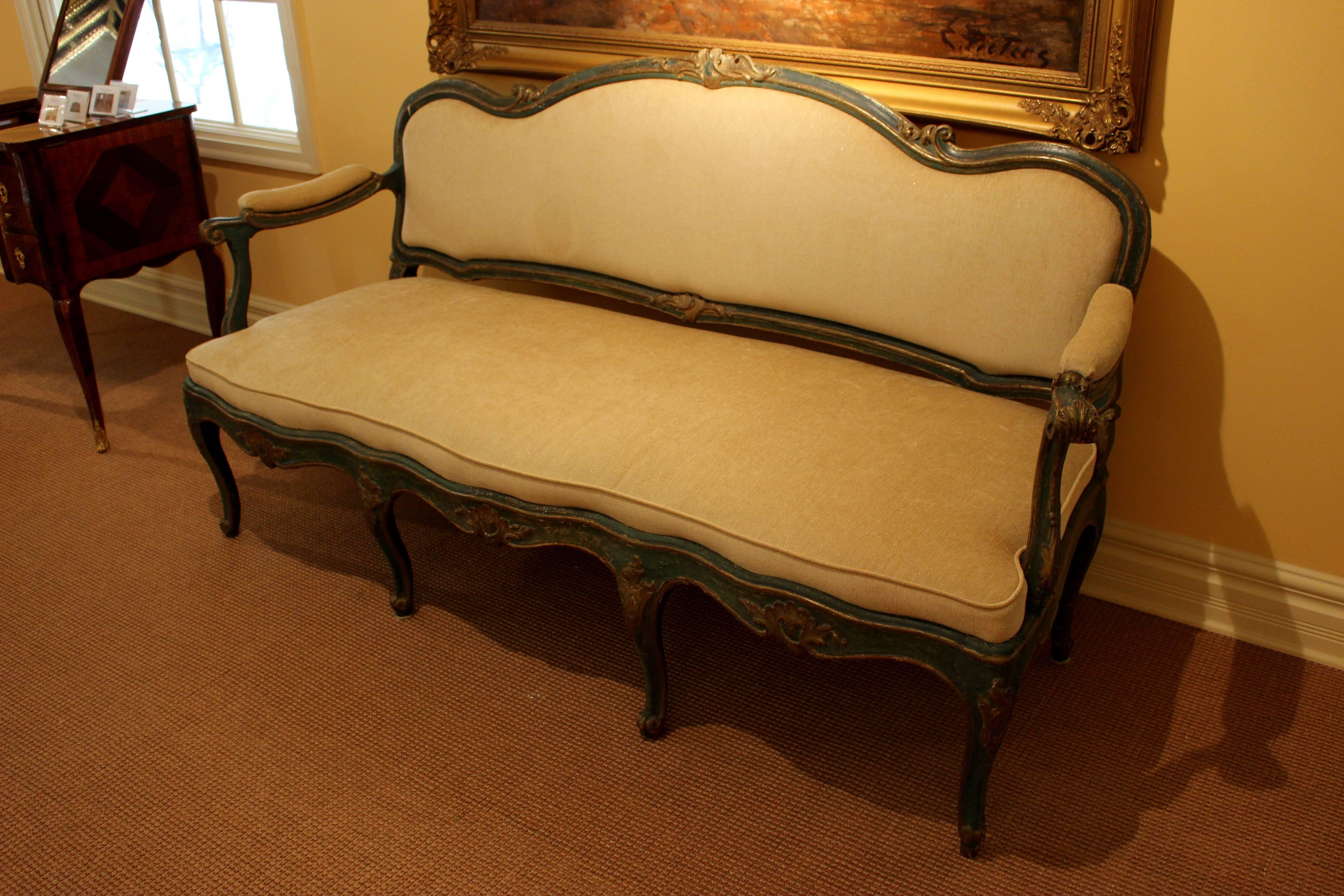 A North Italian green-painted and parcel-gilt settee from the mid-18th century. The serpentine padded backrest displays foliate scrolls on the top rail flanked by two scrolls above a serpentine padded seat. Down-scrolled padded armrests flank the