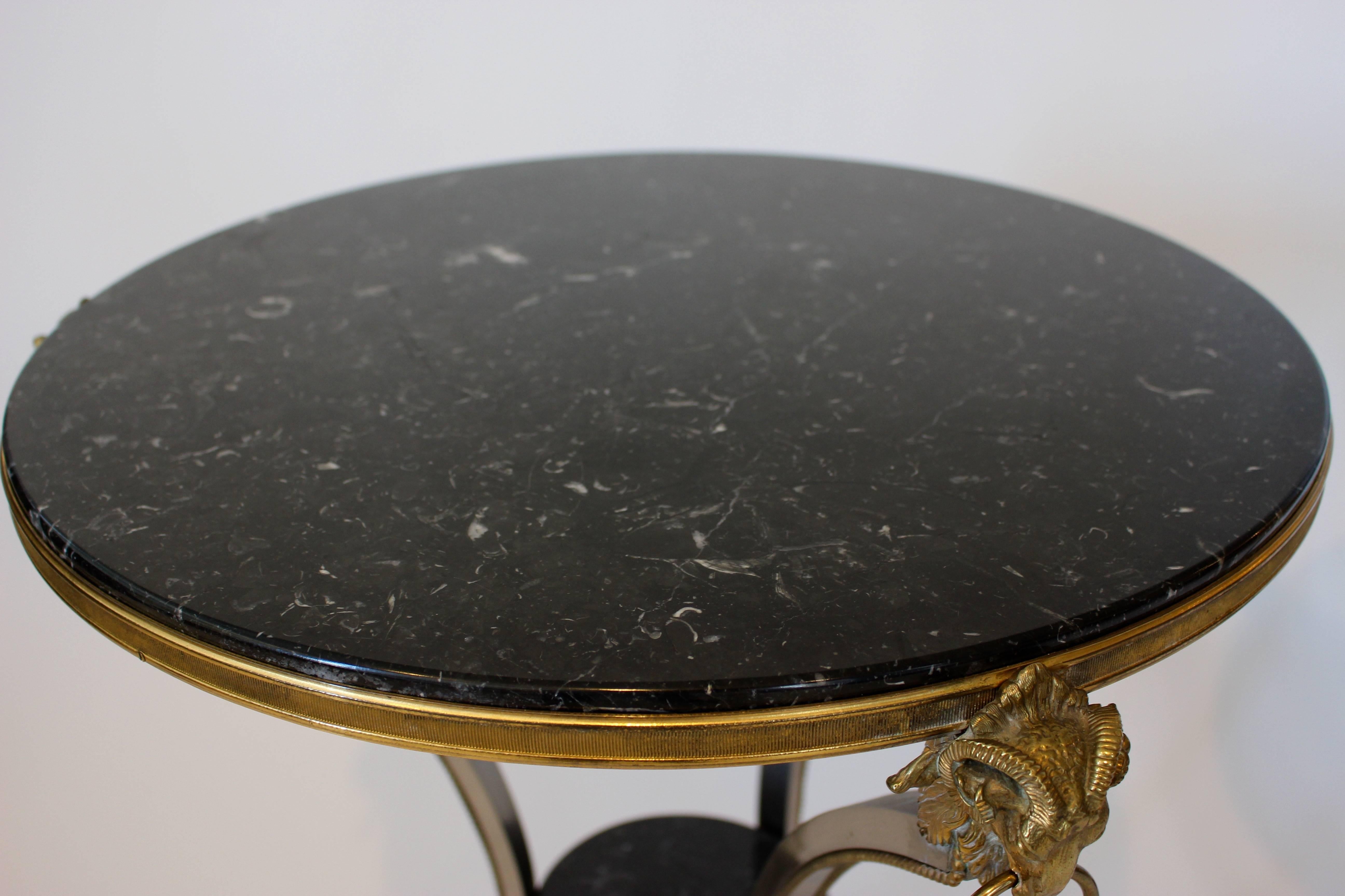 A most elegant associated pair of US ormolu, metal and marble guéridon tables from the 21st century. The tables present a French circular variegated black marble top within a reeded ormolu border. Each piece rests on three inward curved metal legs