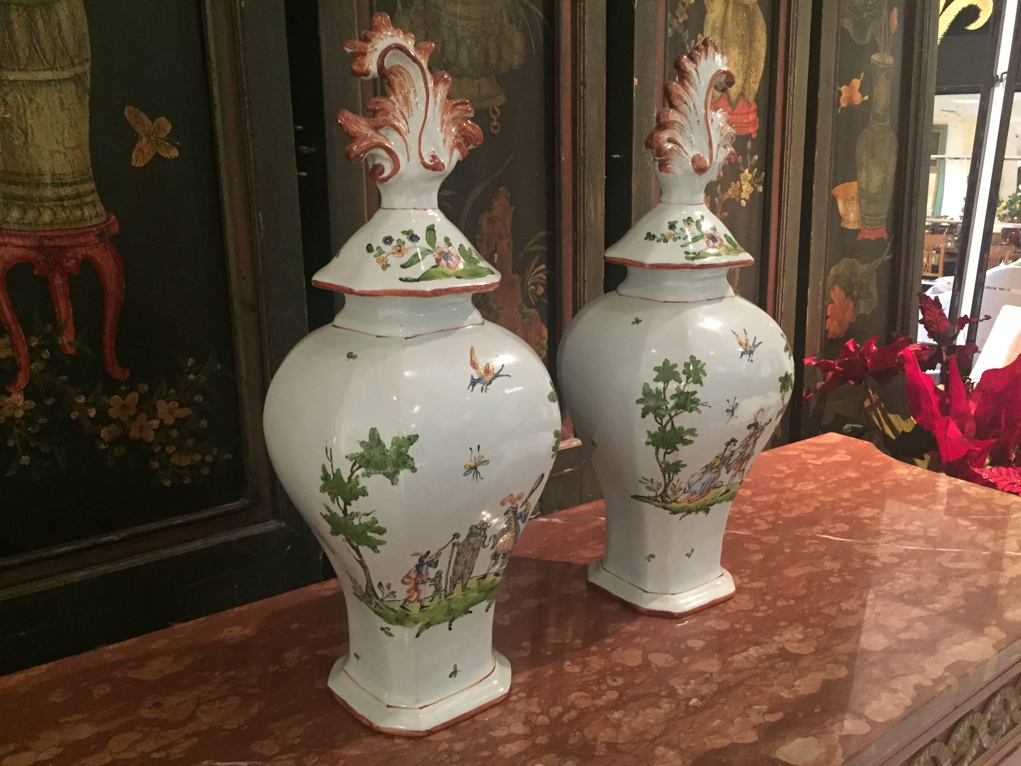 A matching pair of 19th century Italian faience covered vases of flattened baluster form, hand-painted with figures from the Commedia dell'arte. The main body of each piece features a scene with naively painted figures in landscapes on a white