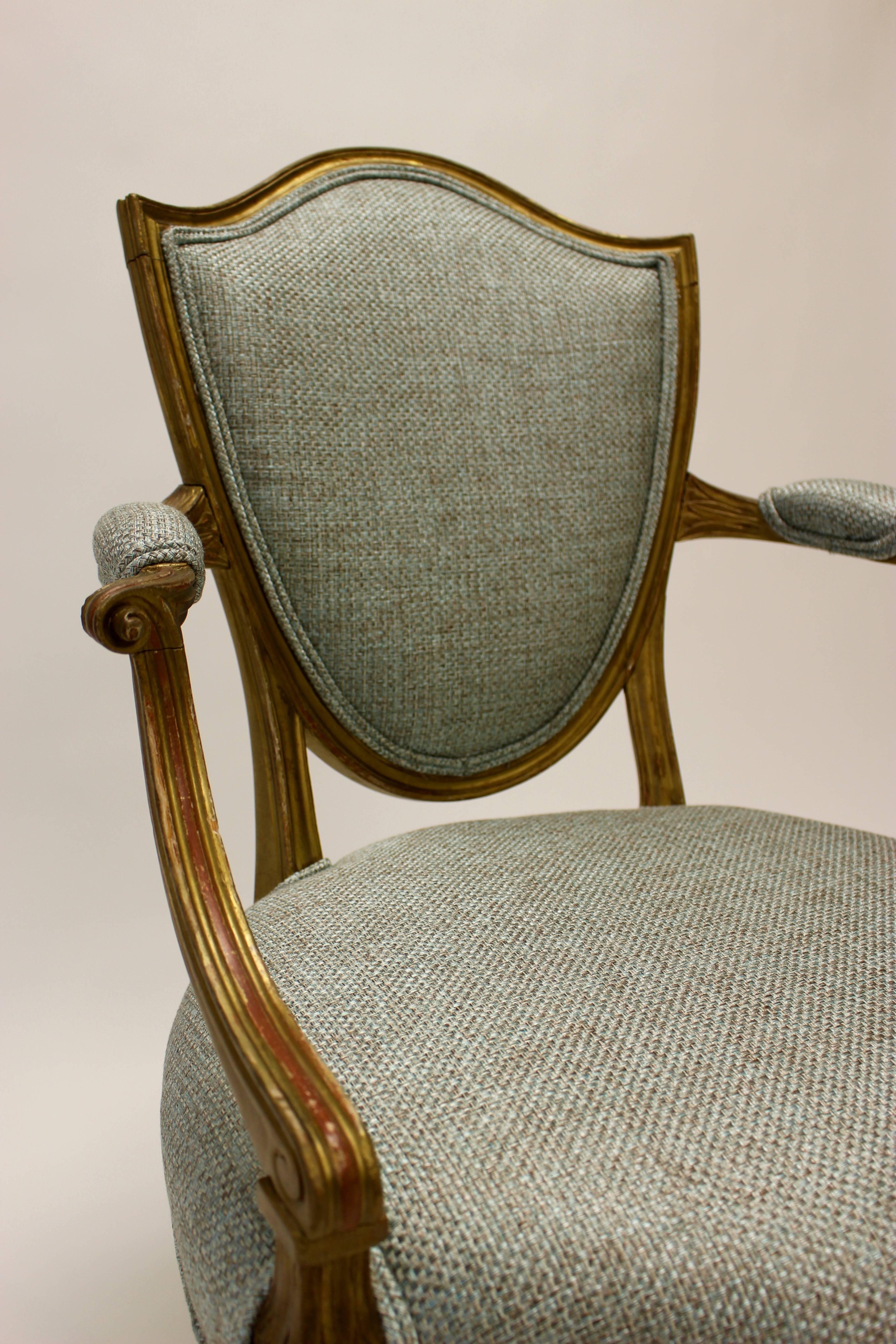 An English 19th century parcel-gilt shield-back chair with open arms in the Hepplewhite style and new upholstery. Featuring a characteristic molded shield-shaped padded back over a round and serpentine padded seat, the chair is upholstered with a