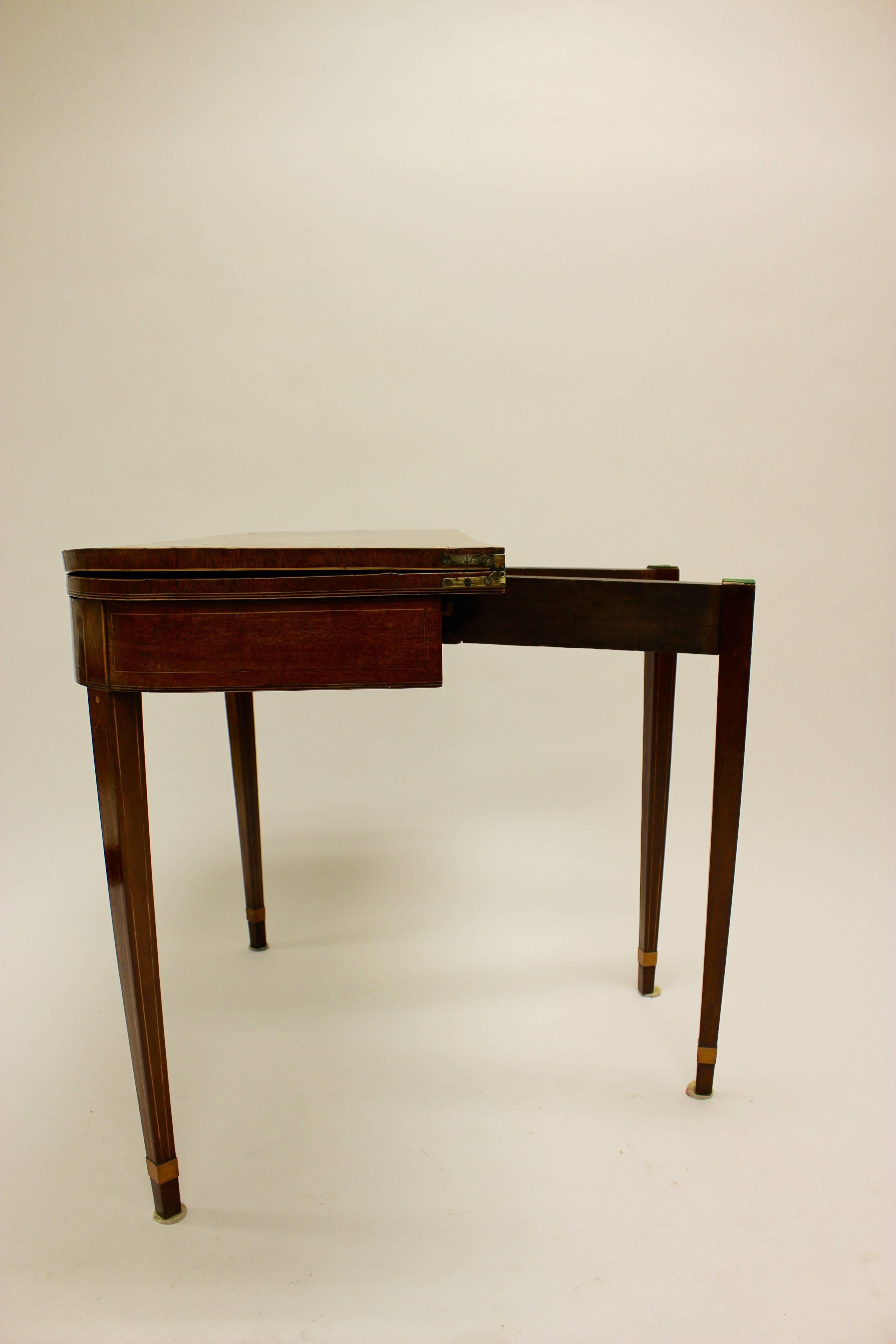 A George III inlaid light-toned mahogany and satinwood card table in the manner of Mayhew and Ince. The D-form cross banded fold-over top opens to reveal a later baize lined playing surface, above a breakfront frieze decorated with cross-banded