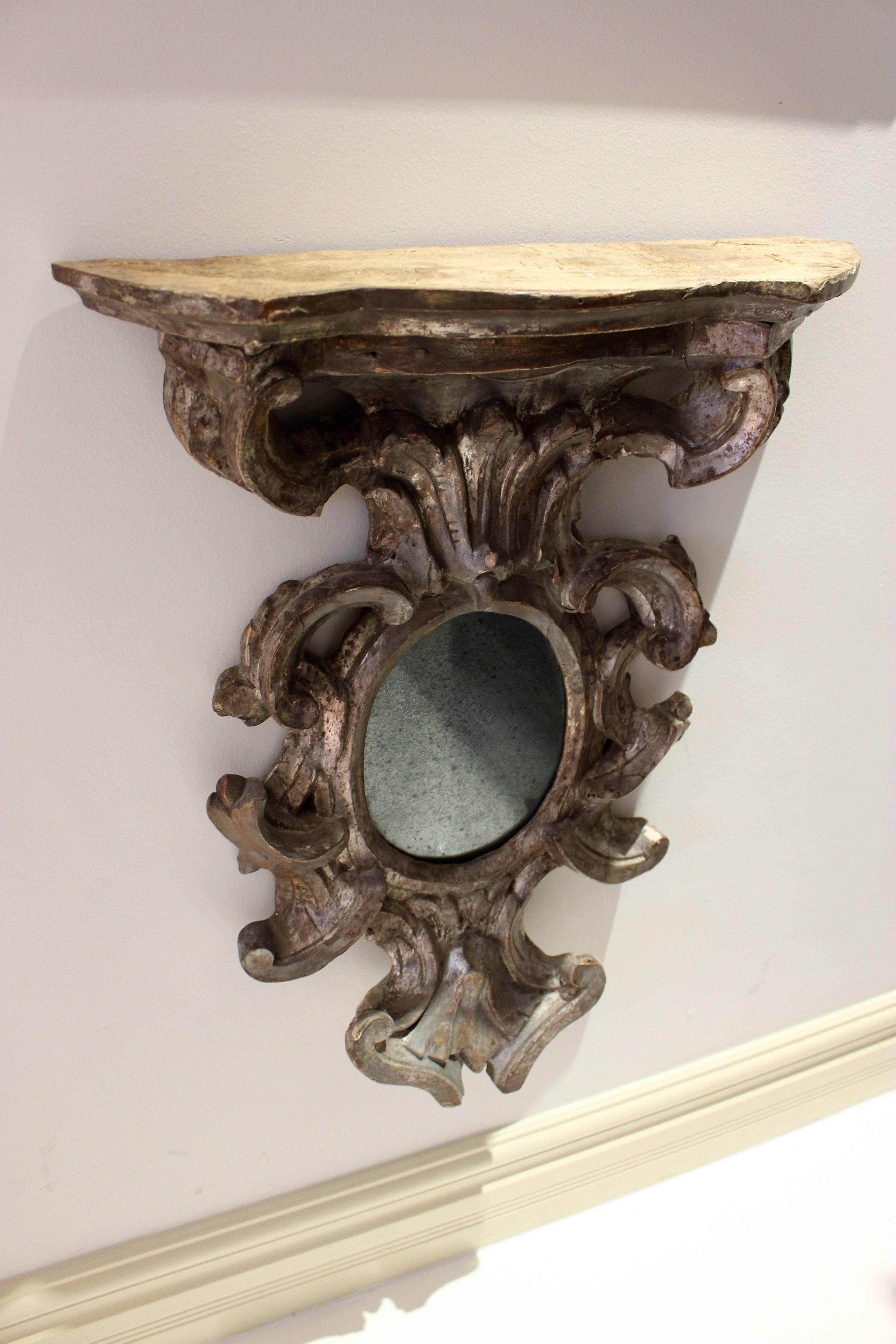 A petite French Baroque style wall bracket with mirror made of carved and silvered wood from the 19th century. The scalloped top rests on a scrolling leaf designed support above a small oval-shaped mirror. Unusually placed below the shelf, the