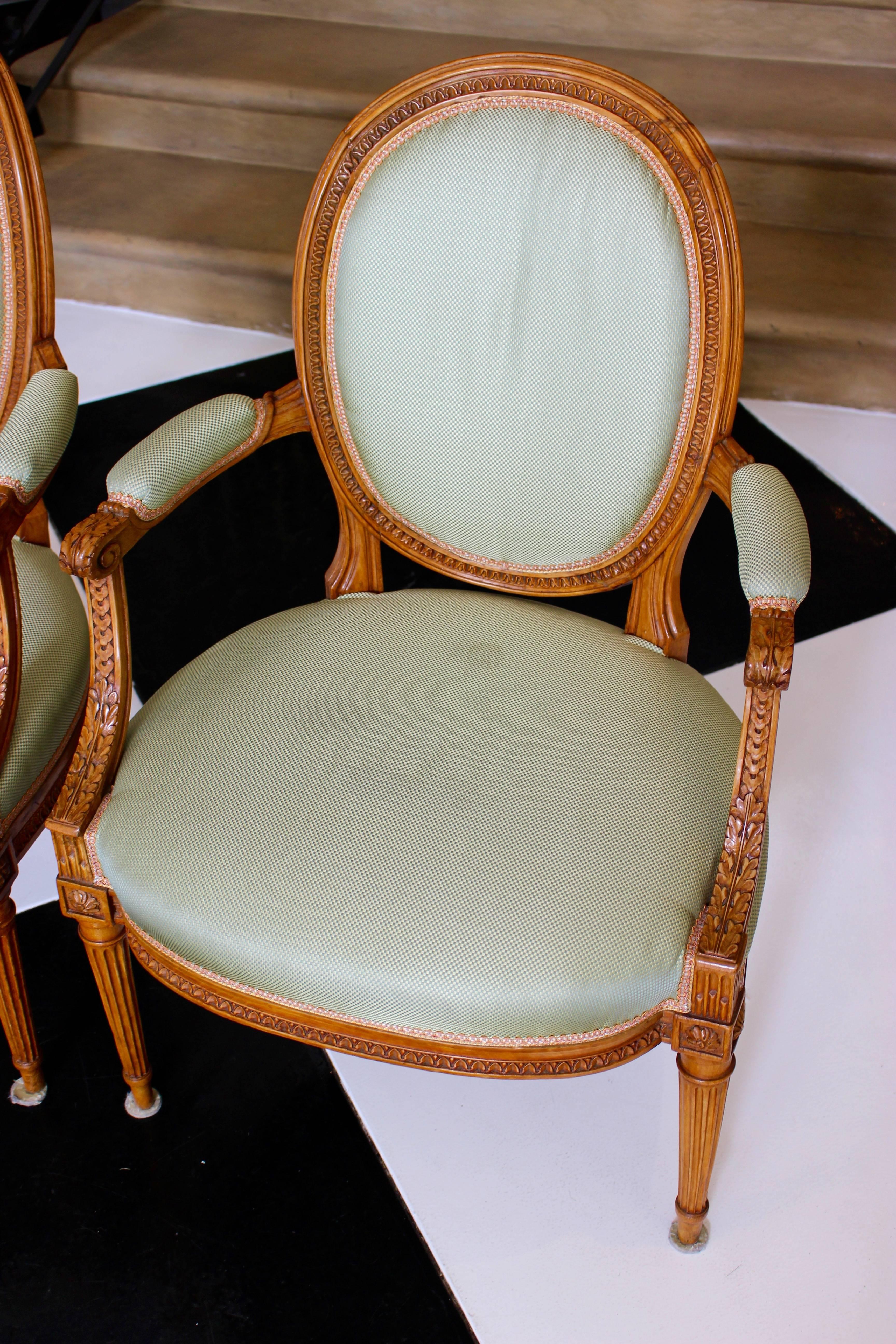 A pair of French Louis XVI style beechwood cabriolets fauteuils with oval medallion backs, scrolled arms, fluted legs and green silk upholstery. Each of this pair of French Louis XVI style armchairs features an elegant oval-shaped molded back,
