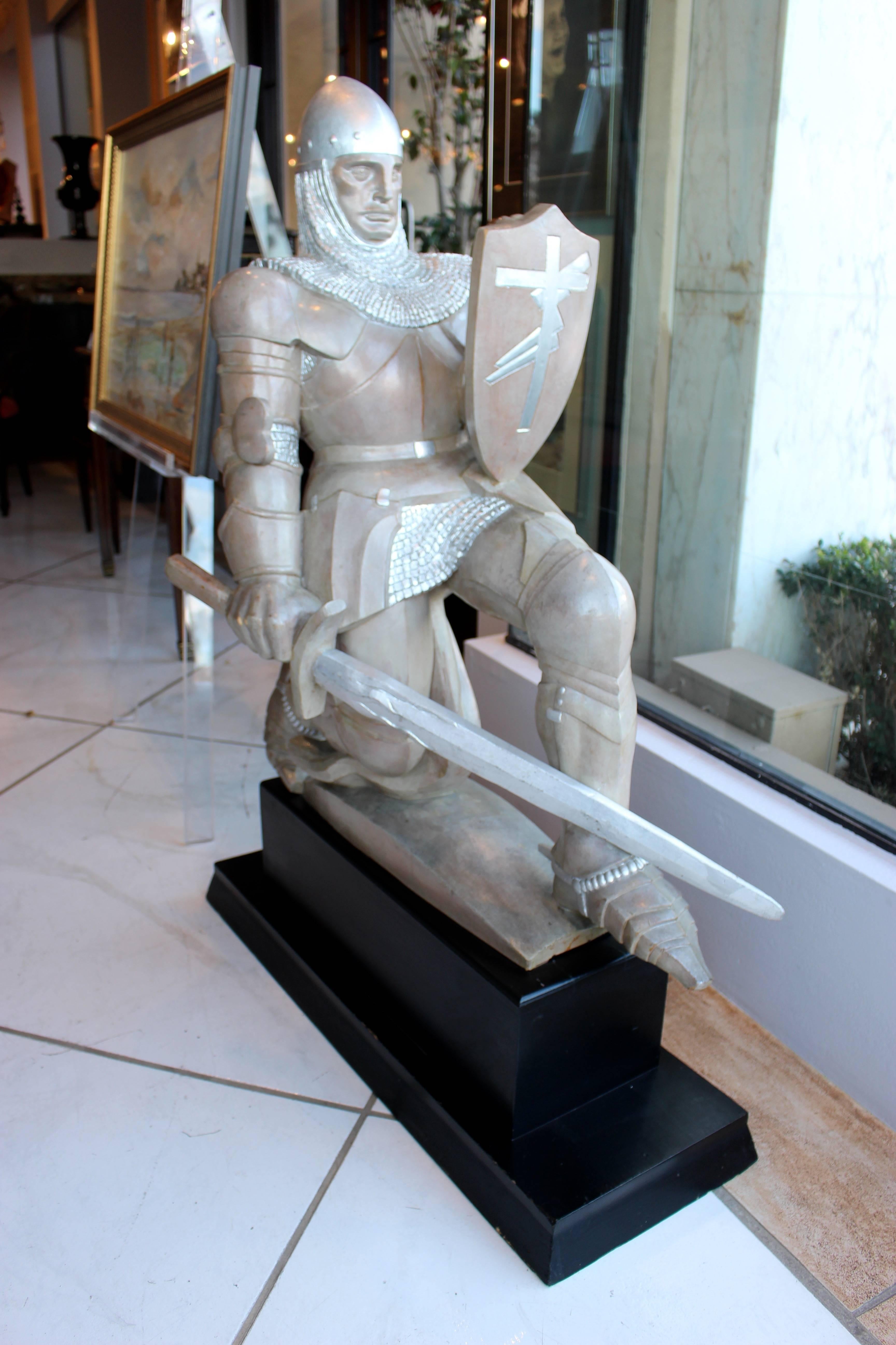 A carved wooden sculpture of a knight crusader from the mid-20th century painted in a silver color with brown undertone. The knight is kneeling on a later rectangular black wooden stepped pedestal, holding his sword horizontally while wielding a