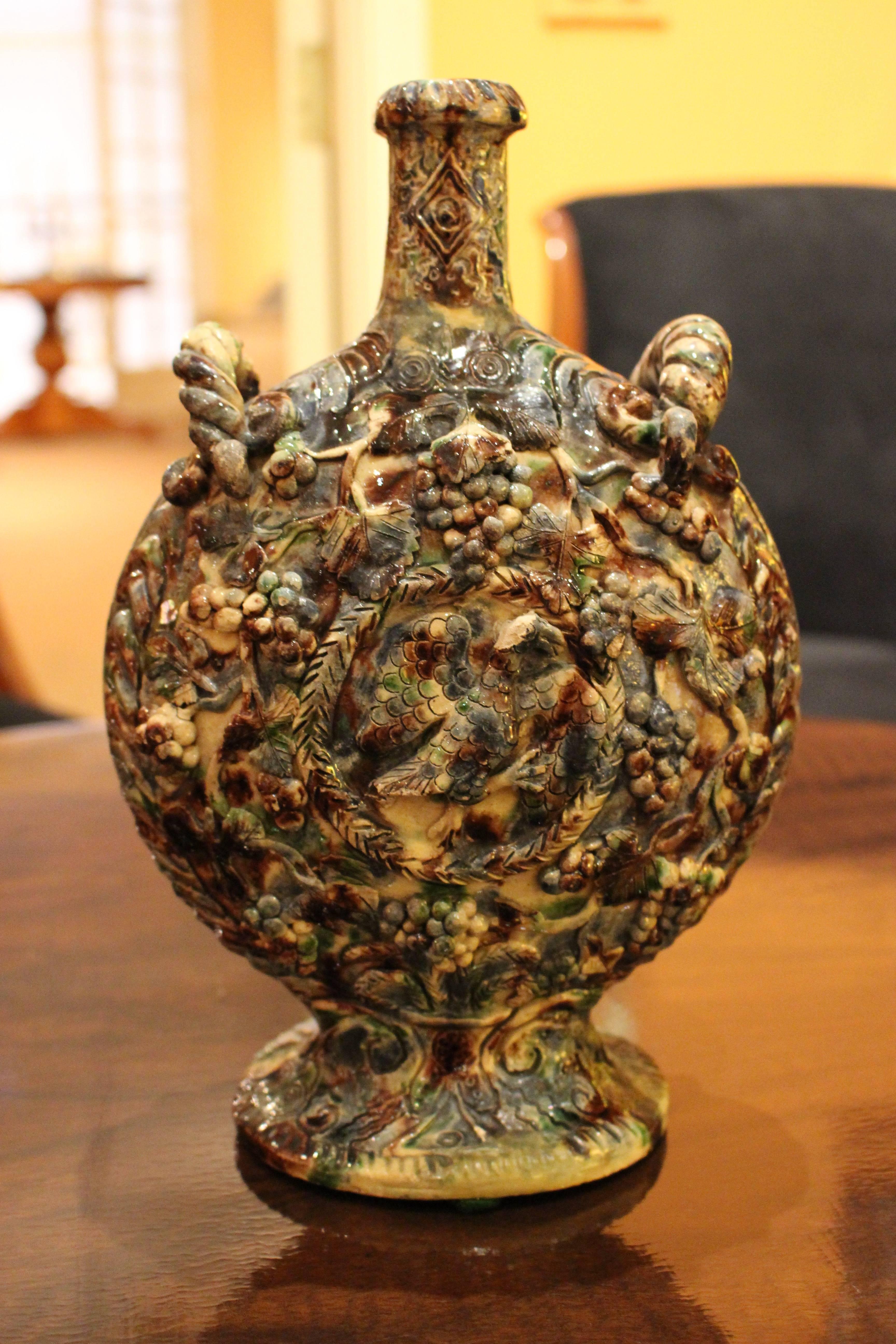 A rustic Italian faience vase molded in relief from the 19th century. The body of flattened circular form is topped by a tall cylindrical neck with a flaring rim flanked by rope-like handles. Molded in relief overall, each side is decorated with the
