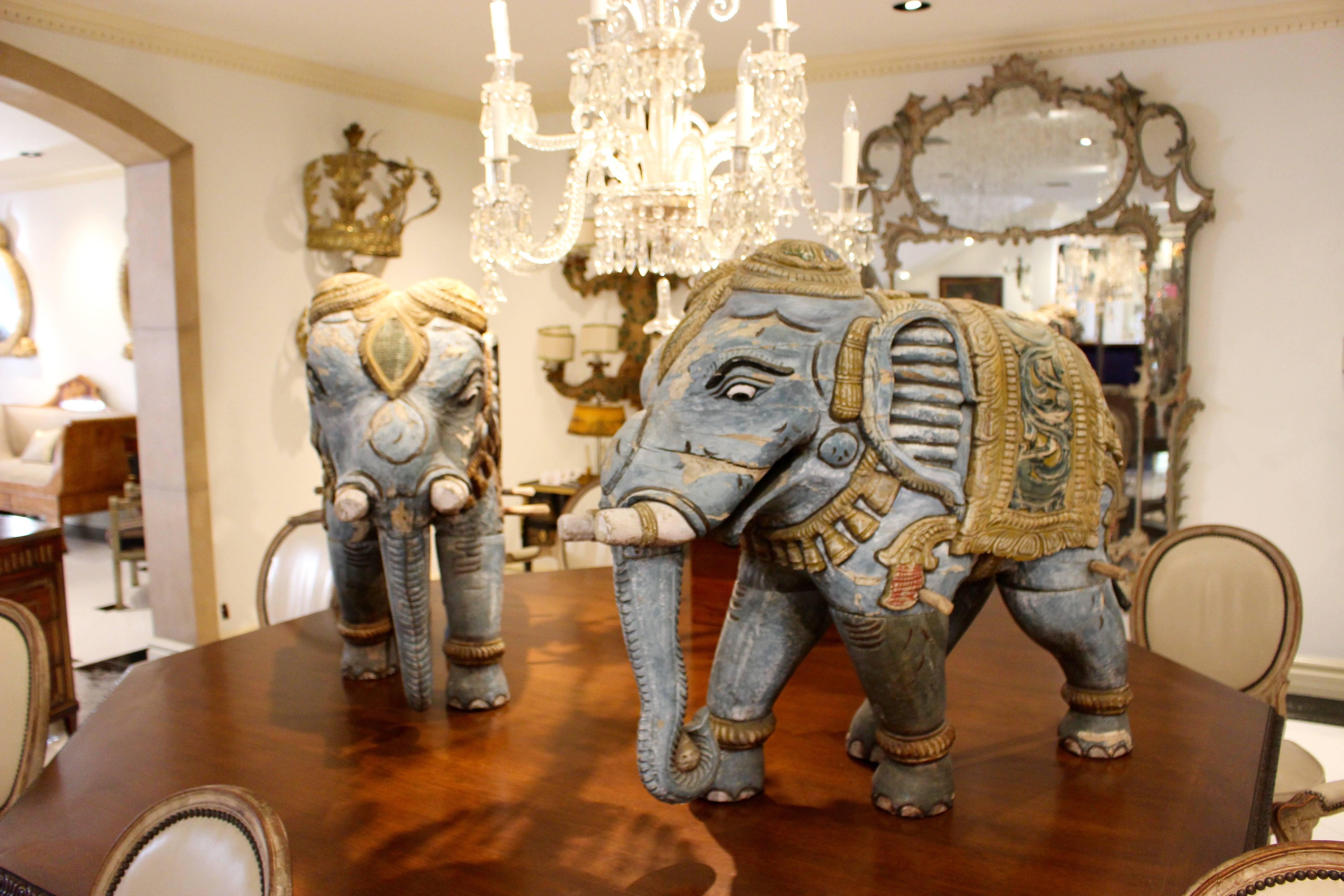 An exotic pair of striding wooden elephants sculptures carved and painted with exquisite detail. Wearing trinkets on the legs and tusks along with a large ceremonial necklace, each figure is caparisoned with an ornate yellow howdah blanket