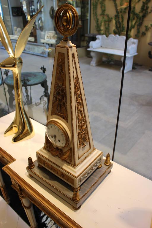 A French white marble mantel clock of obelisk form with ormolu decorations in Louis XVI style. The enamel dial with Arabic numerals and pierced gilt hands is enclosed in a white marble obelisk case surmounted by a gilt moving armillary sphere finial