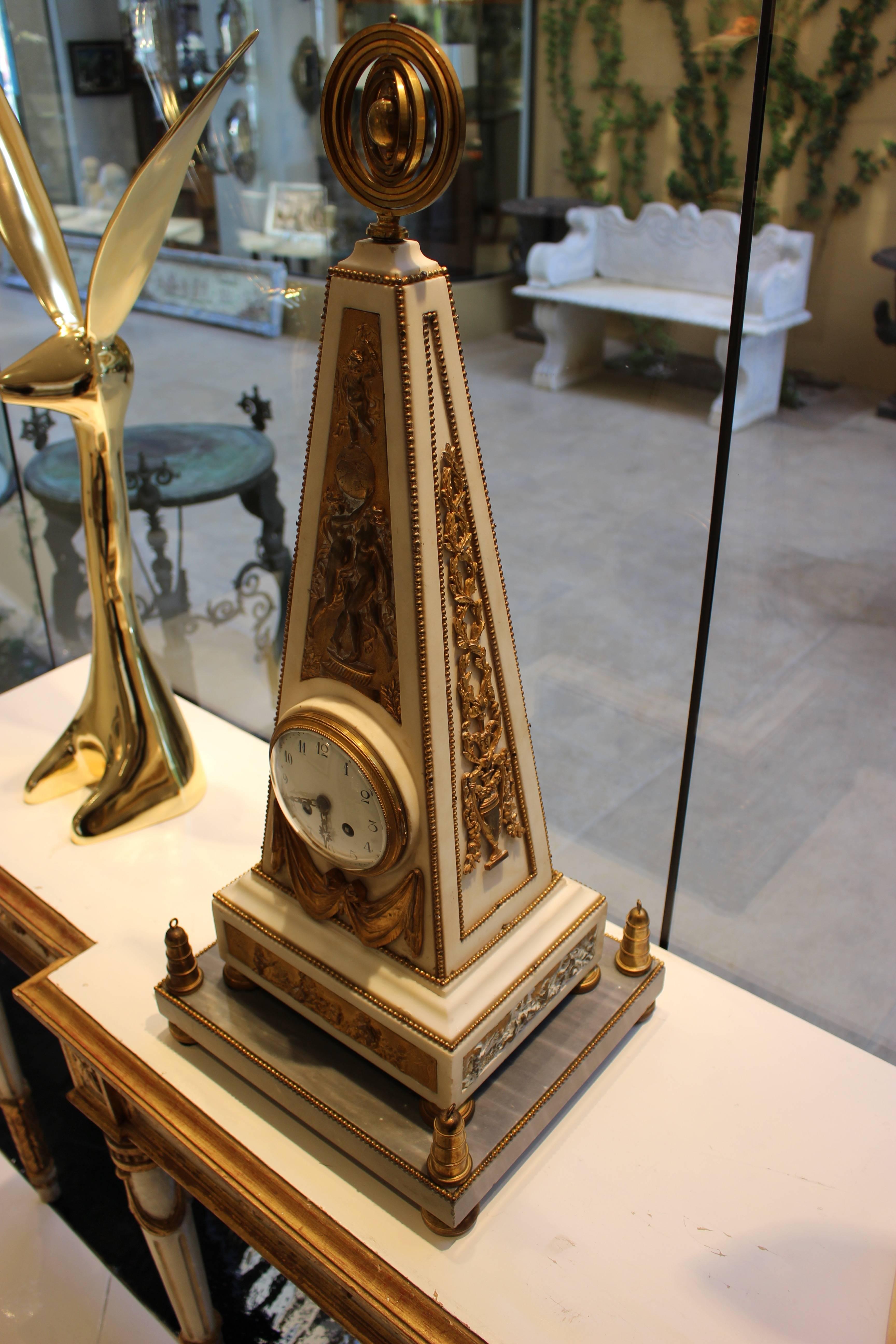Pendule de cheminée en marbre blanc français de forme obélisque avec décorations en bronze doré de style Louis XVI. Le cadran émaillé avec chiffres arabes et aiguilles dorées percées est enfermé dans un boîtier obélisque en marbre blanc surmonté