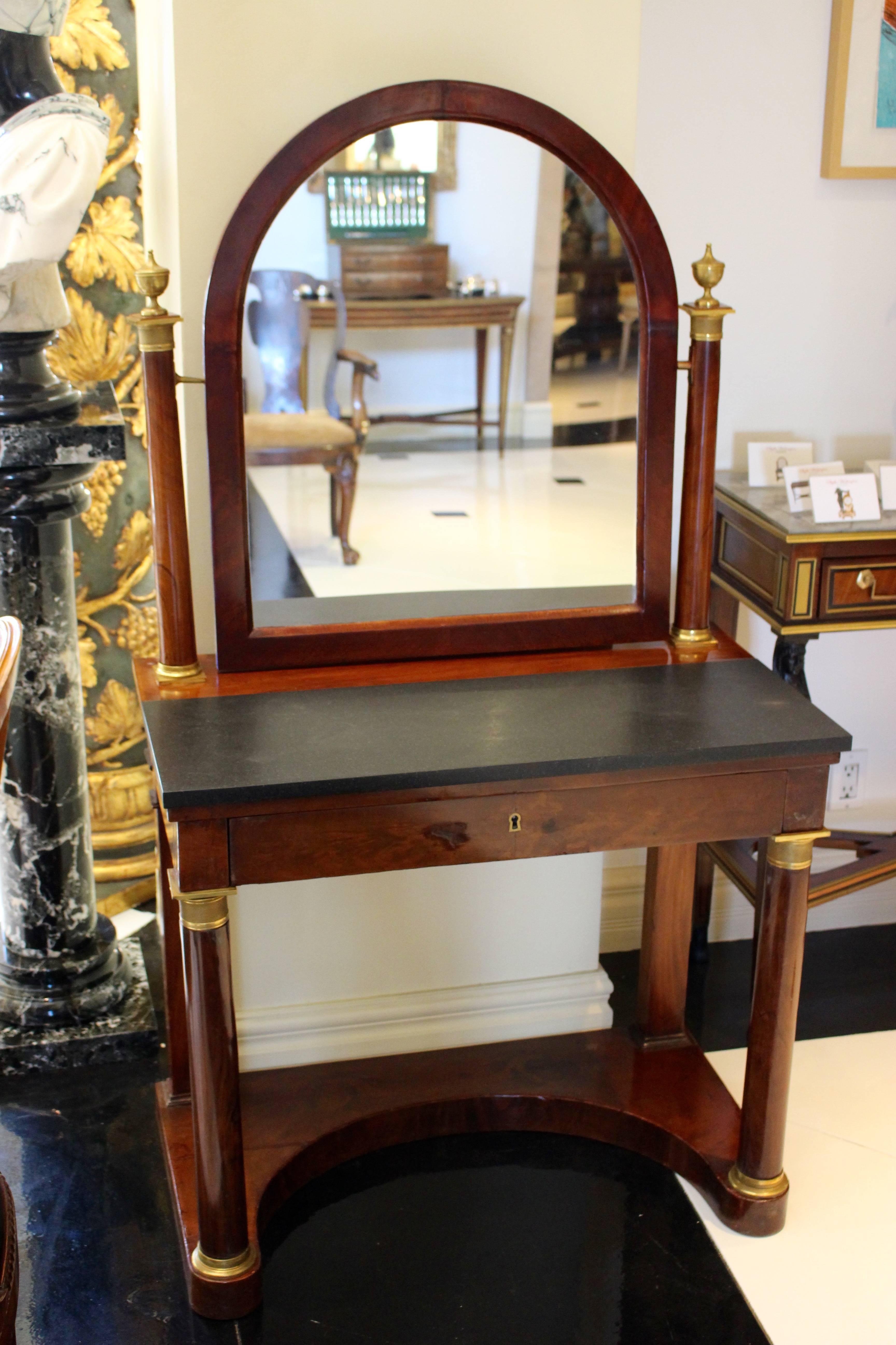 A French Empire period ormolu-mounted mahogany psyché dressing table from the early 19th century in neoclassical style. The rectangular black marble top is surmounted by an arched tilt mirror flanked by mahogany columns capped with urn finials above