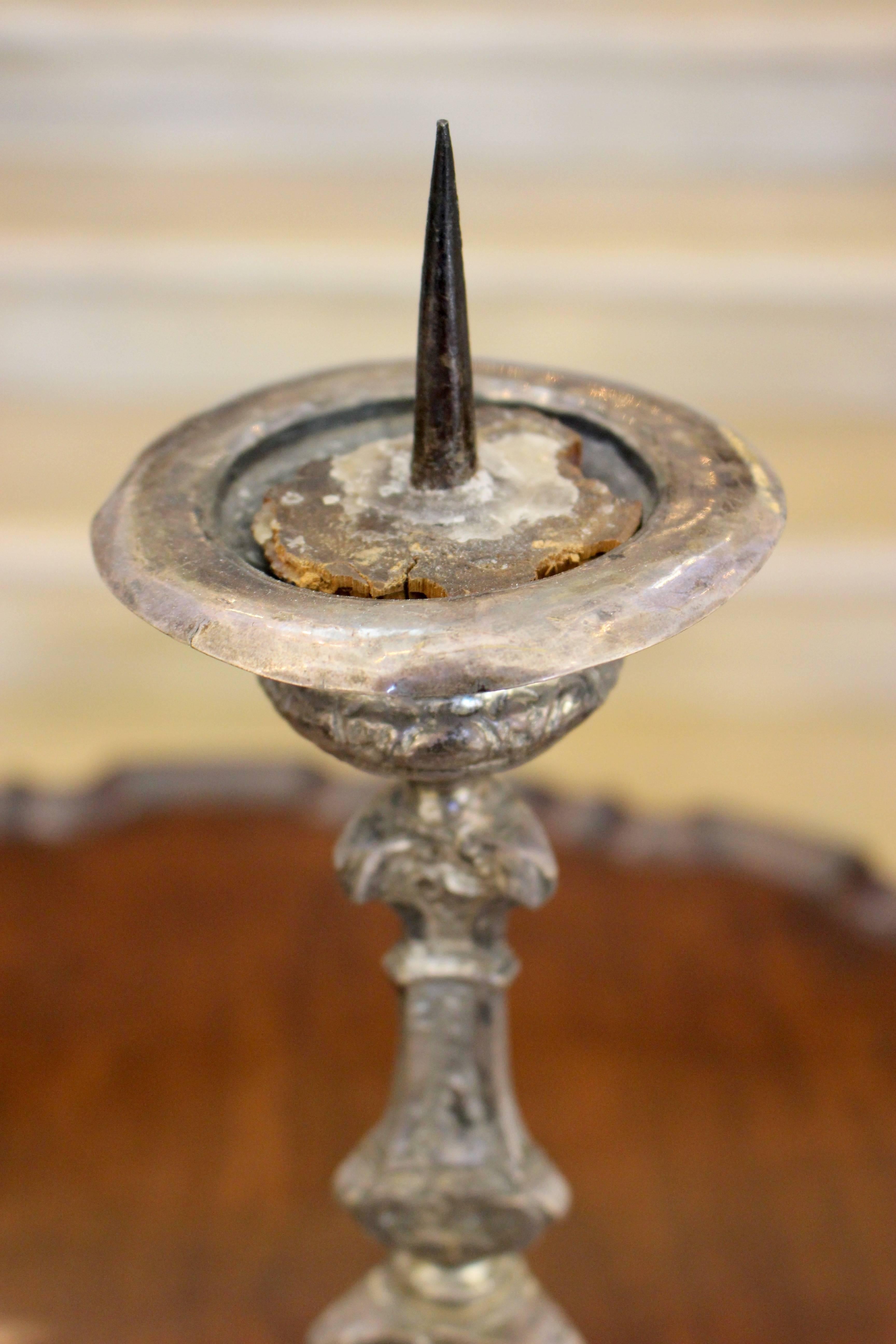 A pair of decorative matching silvered brass pricket candlesticks resting on a triform base with embossed Baroque foliate and scrolls motifs. Each presents a multi-baluster stem supporting an elegant urn-shaped drip pan from which tall prickets