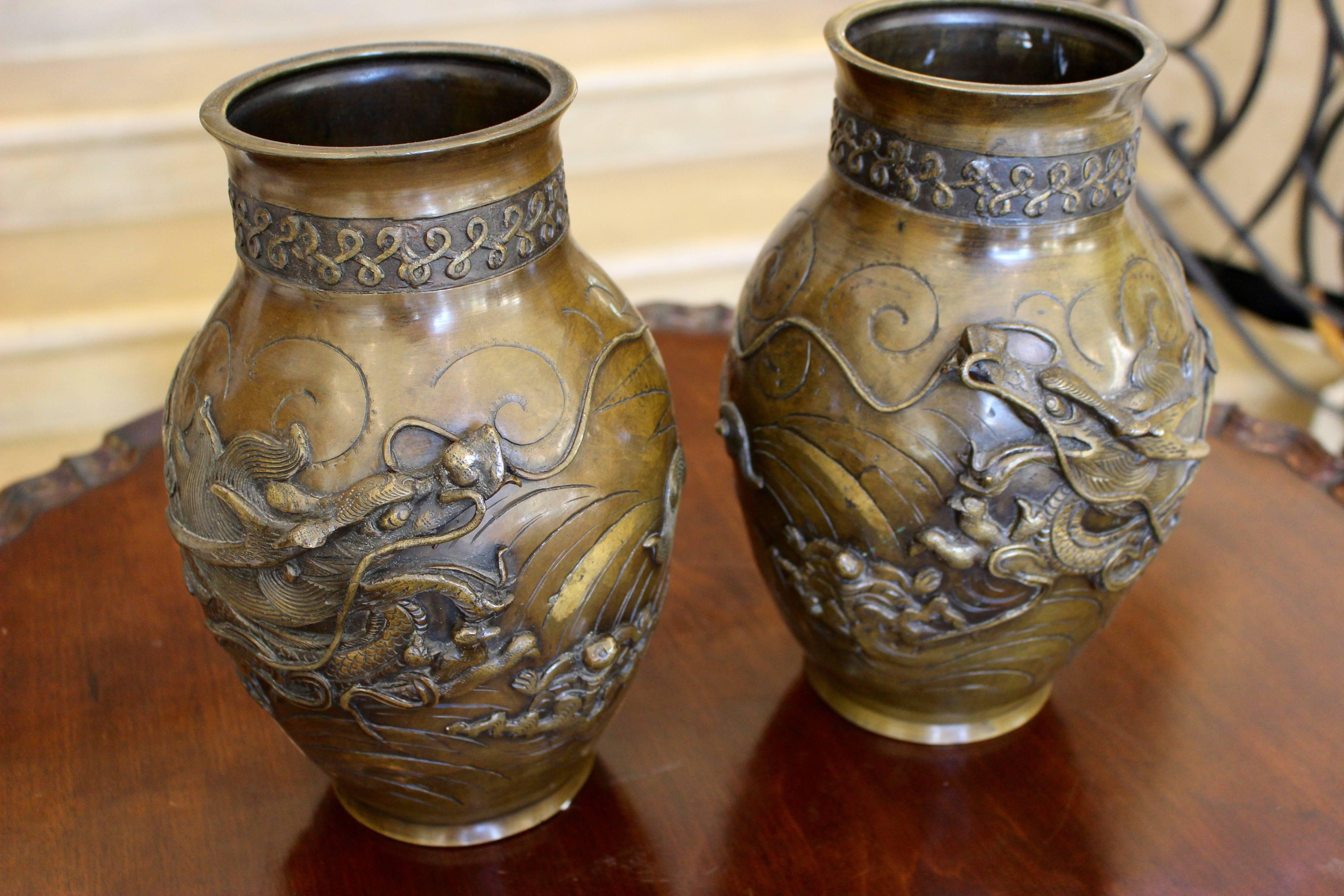 A pair of matching Japanese bronze vases from the 20th century finely cast with dragon figures. A high relief cast decoration of a three-clawed dragon is wrapped around the body of each vase, swimming in an aquatic environment symbolized by curved
