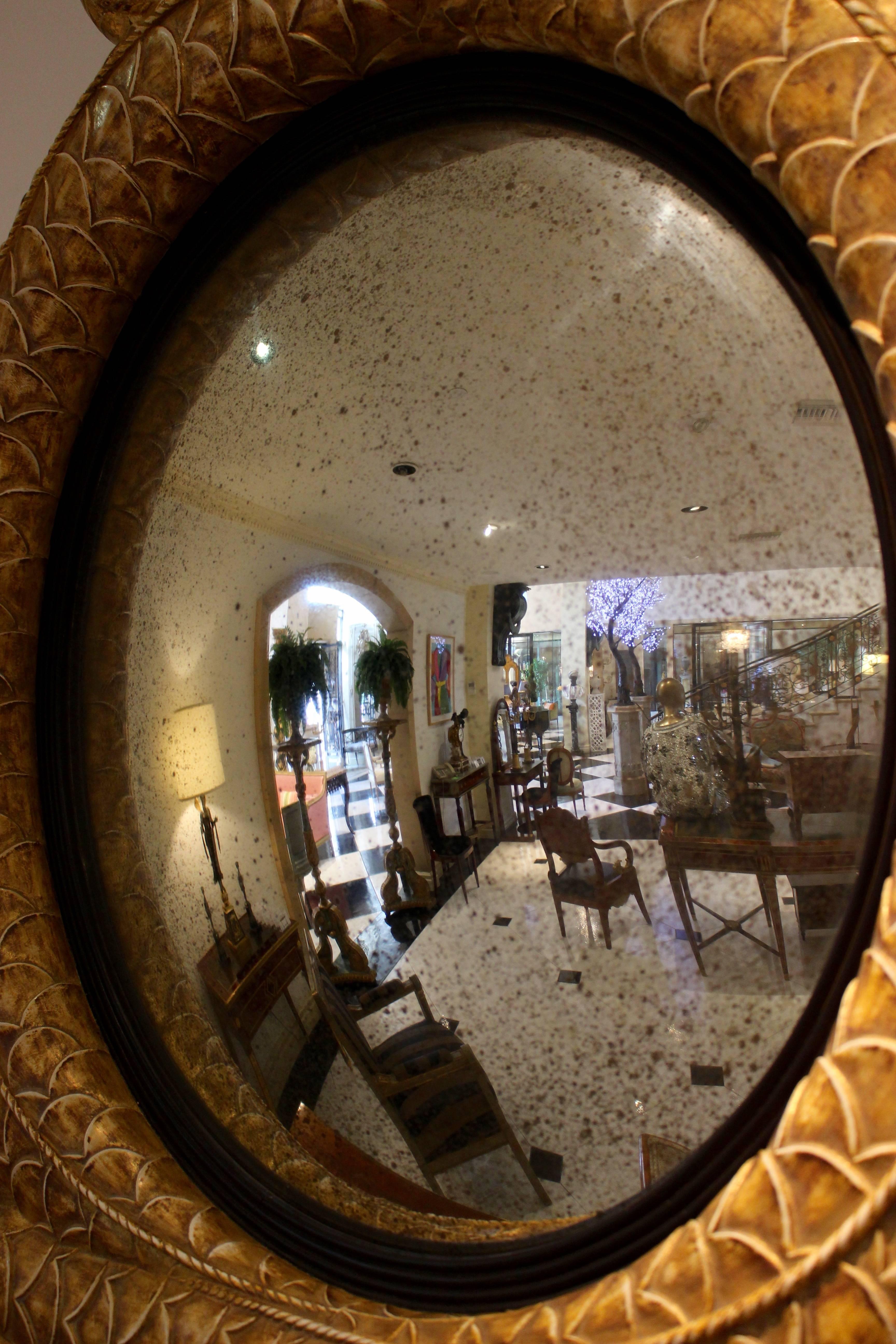A pair of Regency style giltwood and parcel-ebonized convex dolphin mirrors from the 21st century. Each mirror presents a distressed convex round mirror plate within an ebonized inner frame. The larger giltwood frame is shaped as two stylized