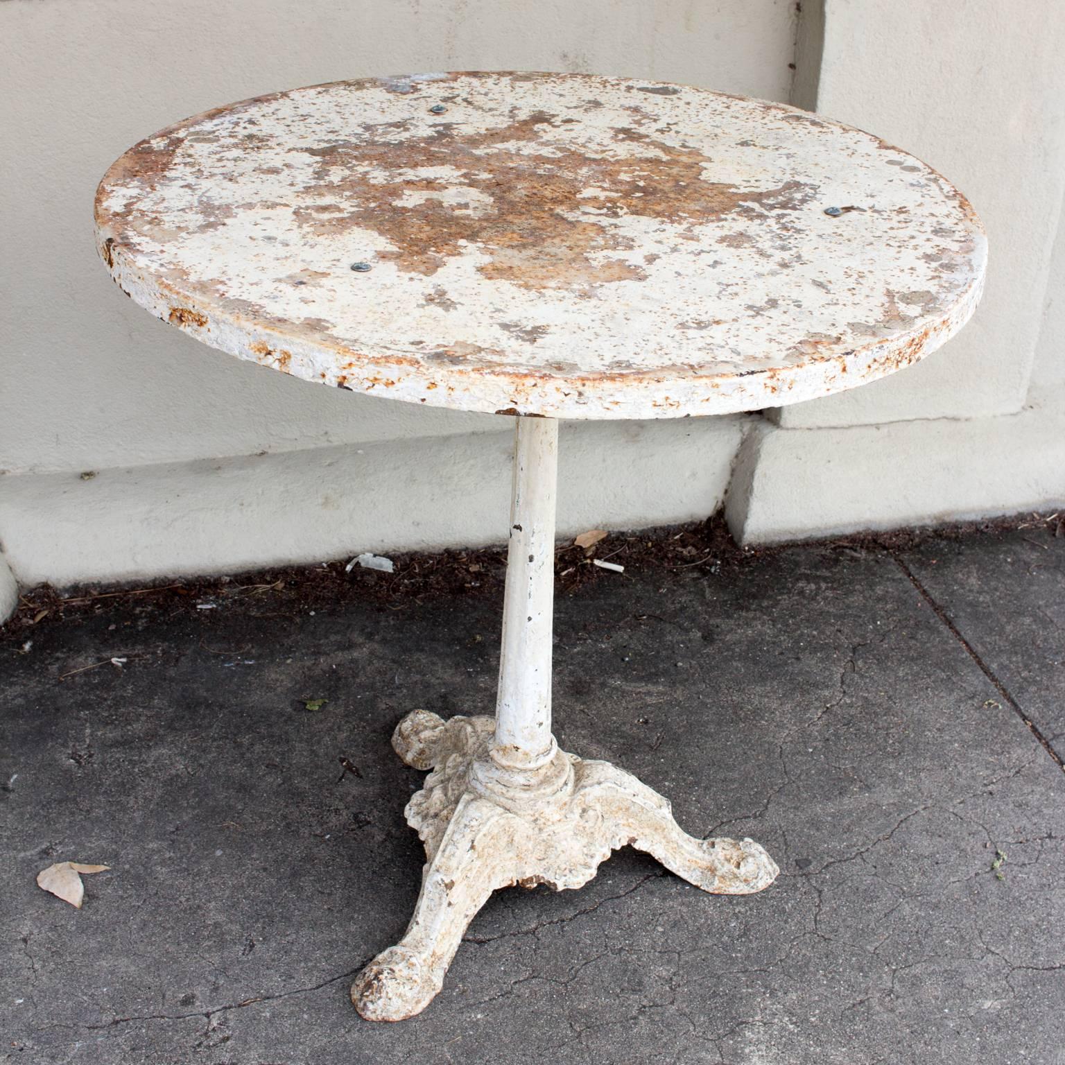 This 1920s bistro table features a cast iron base with a metal top that has been painted a creamy ivory color. The top is secured with three small screws, one of which is missing, but can easily be replaced. This piece has rustic, French charm as