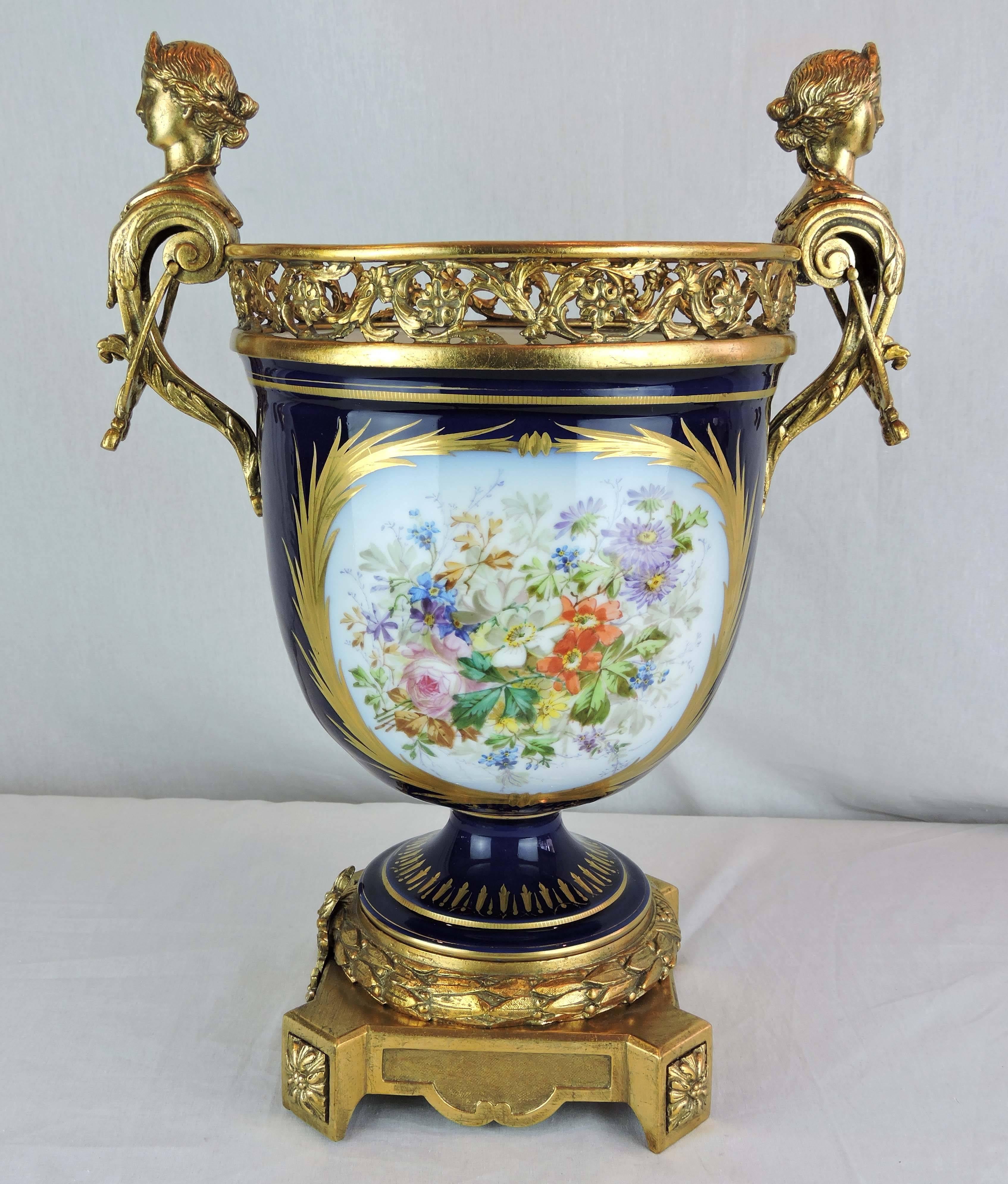 This is an urn-shaped, large French Sèvres Porcelain pedestal jardinière. It is surmounted by a pierced, foliate, gilt bronze gallery framed between a pair of neoclassical figural handles. 

The cobalt blue body is decorated with two, large,