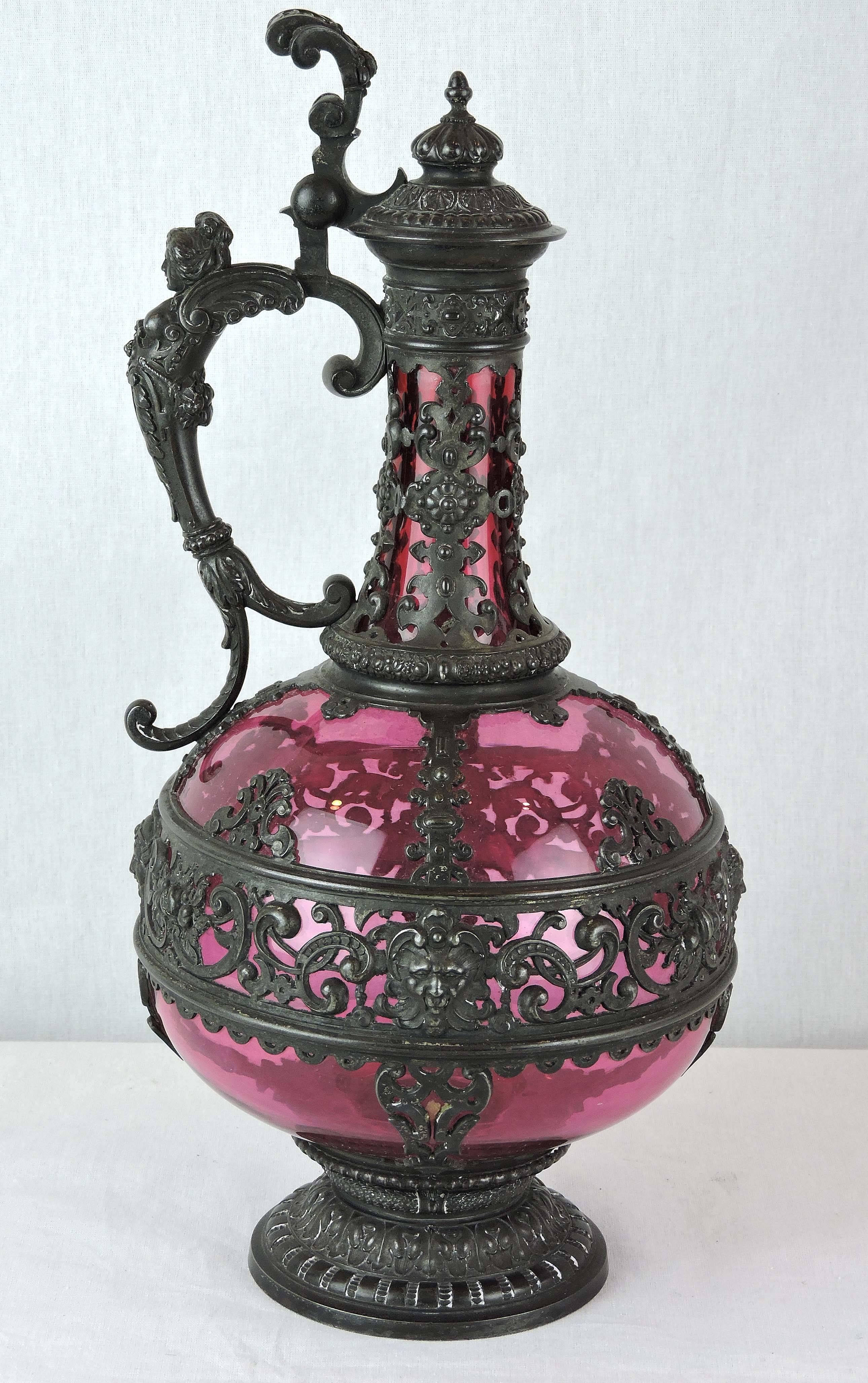 This is a 19th century Jacobean Revival cranberry ewer: The slender, elongated neck joined to a globe-shaped body, mounted within a metalwork frame, topped with a domed hinged lid, with a foliate lever rising above the handle, in the form of a