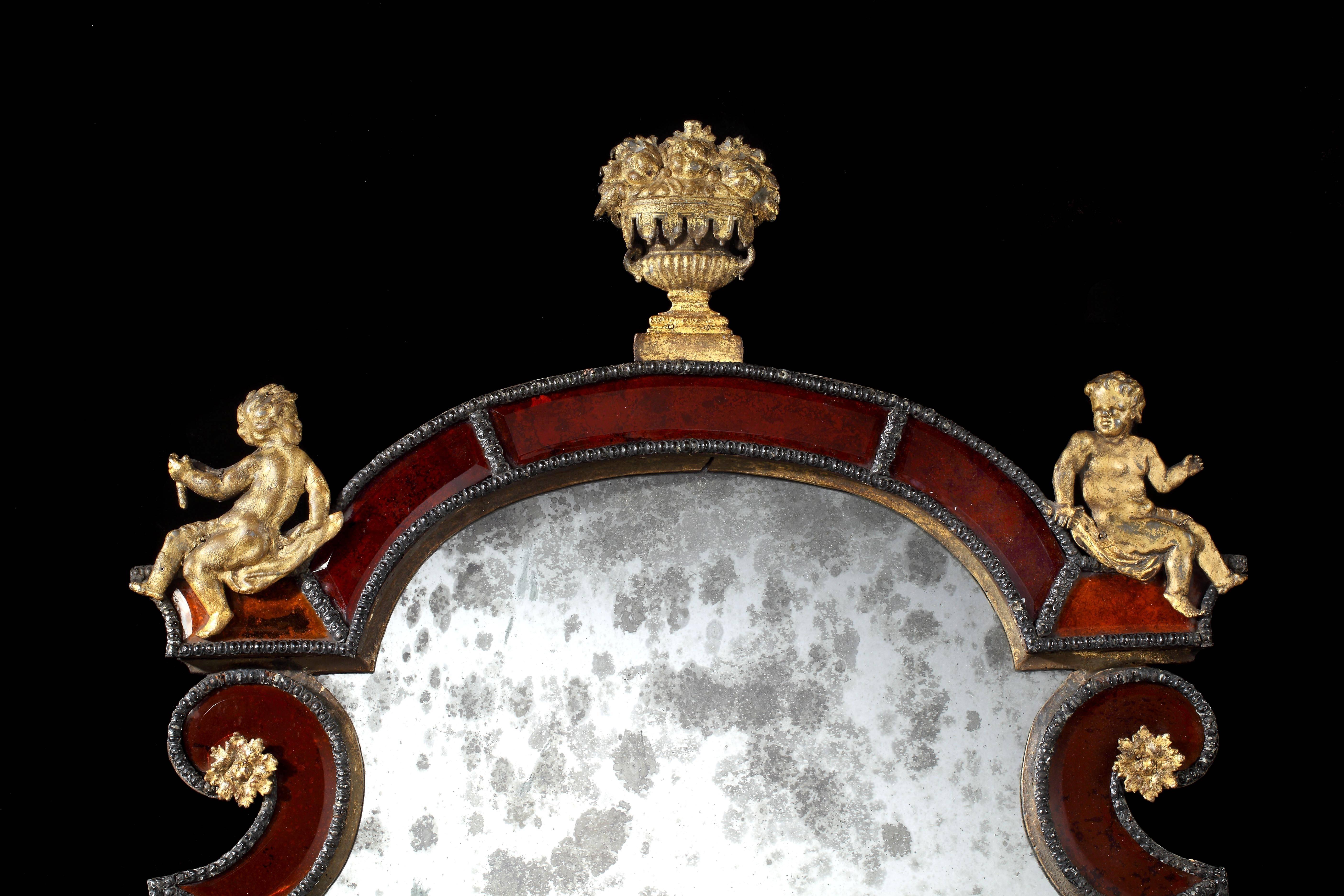 Sweden, circa 1720. Probably by Gustav Precht (1698-1763).

The frame of crimson glass with gilt-lead and giltwood mounts, with flanking putti and central flower basket, retaining the original rectangular plate, the shaped upper plate almost