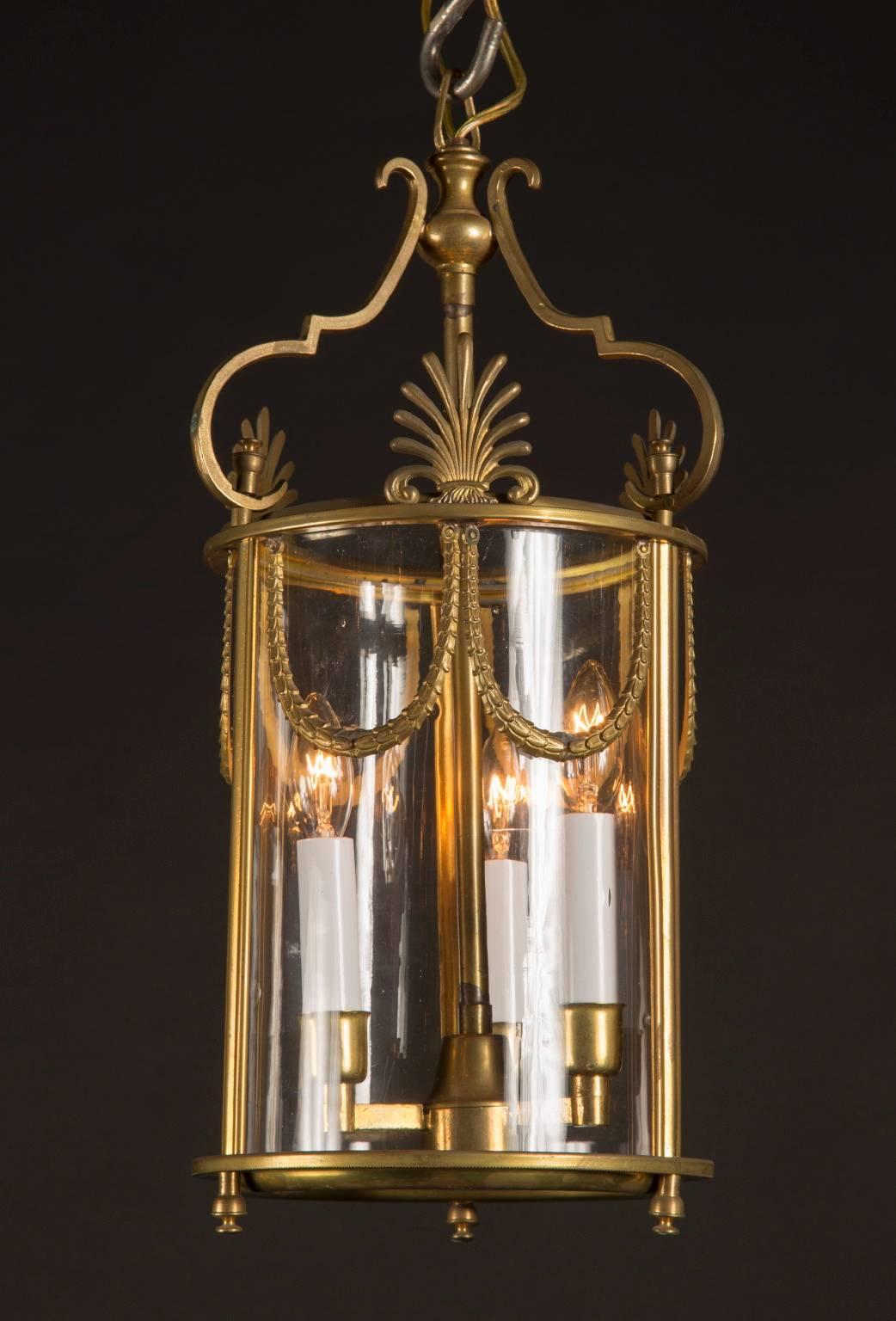 These small cylindrical lanterns are decorated with streams of garlands hanging down from the upper metal rim of the lantern. A trio of palmetto fronds sits atop the metal rim, spaced between three arms that hold the stem of the lantern. The glass