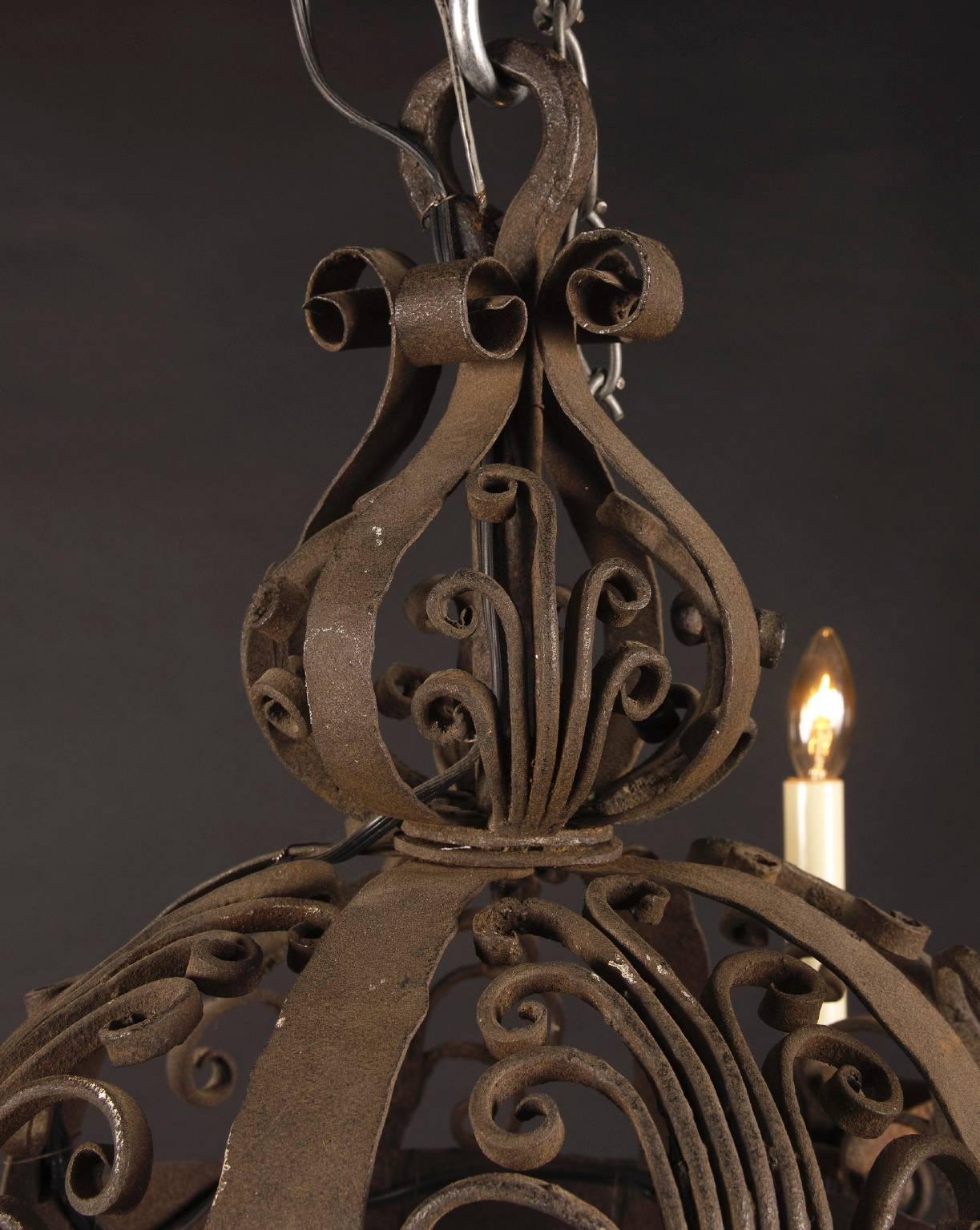 This small 18th century spherical chandelier is made from hand wrought iron circa 1780. The French antique piece features iron bobeches and floriate scroll decorations at top, with a floriate finial. It’s classically medieval in design and draped in